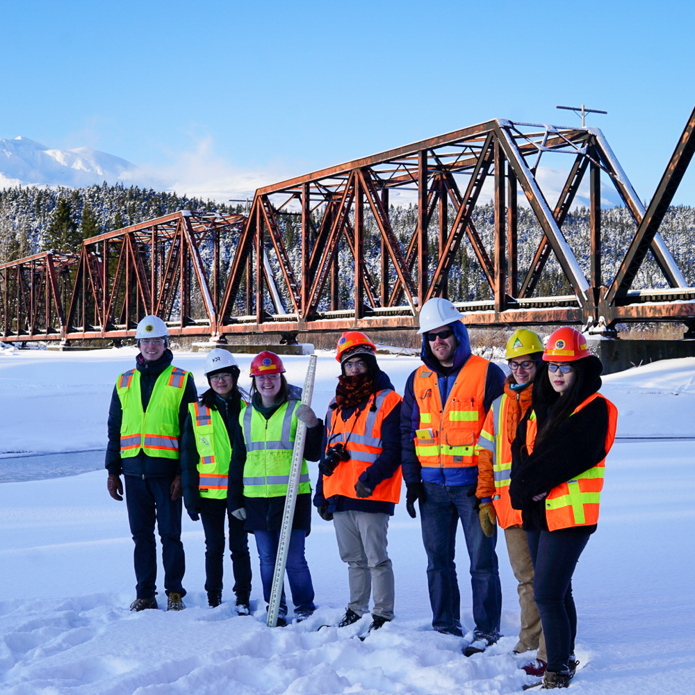 Group of students in reflective vests and hard hats in front of a bridge and mountains