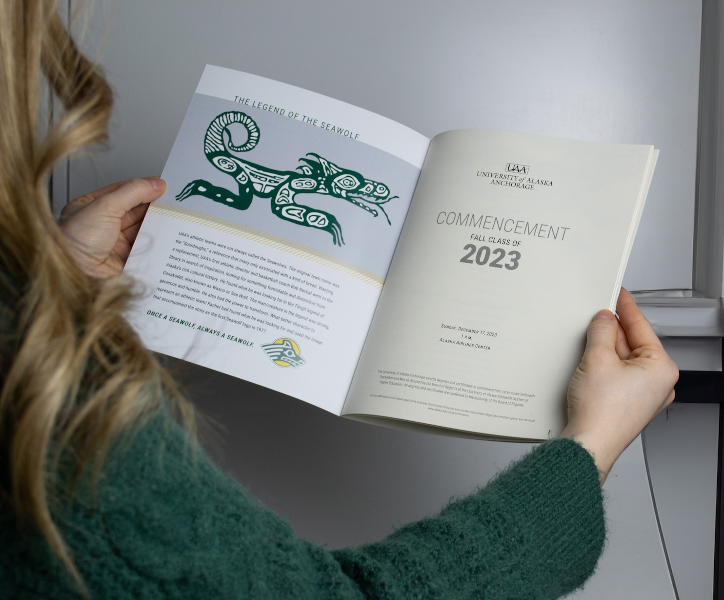 The photo is over the shoulder of a blonde woman in a green sweater. She is holding out a booklet that reads Commencement 2023. She is in a white and grey room under studio lighting.  