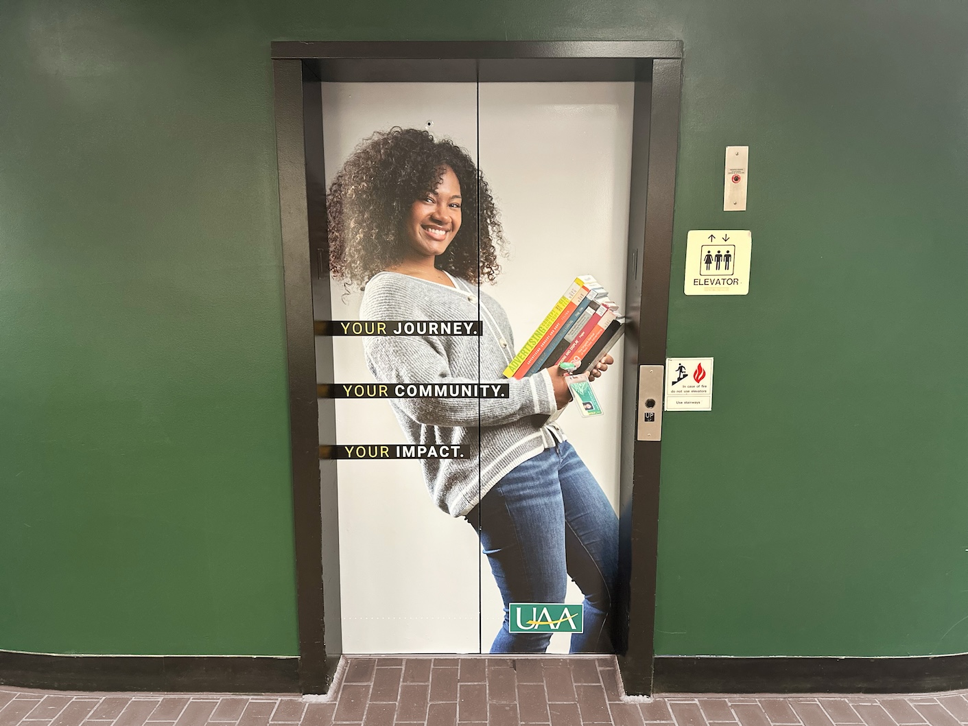 This is an image of an elevator large format graphic. The elevator is located on UAA Campus. The image is of a UAA student, she is holding a stack of books and leaning back. She is smiling and appears happy. She has a UAA ID hanging off the stack of books.