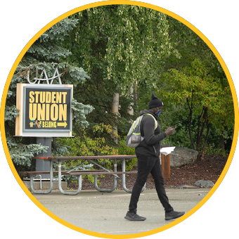 Student with mask on looking at phone, walking outside by sign for Student Union.