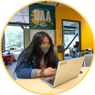 Student wearing mask, working on laptop, with large UAA sign behind her.