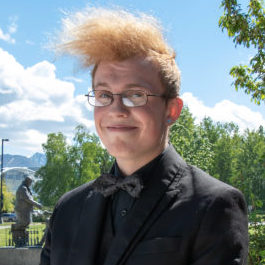 Student, Brett Keene, poses in spring wearing a suit coat and hair blowing in the breeze.