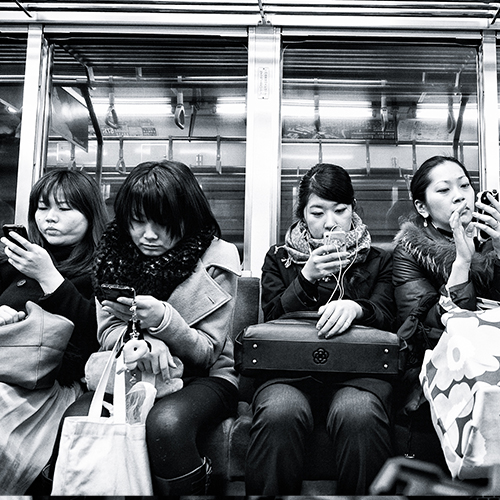 Black and white photo of four women on a train looking at their cell phones.