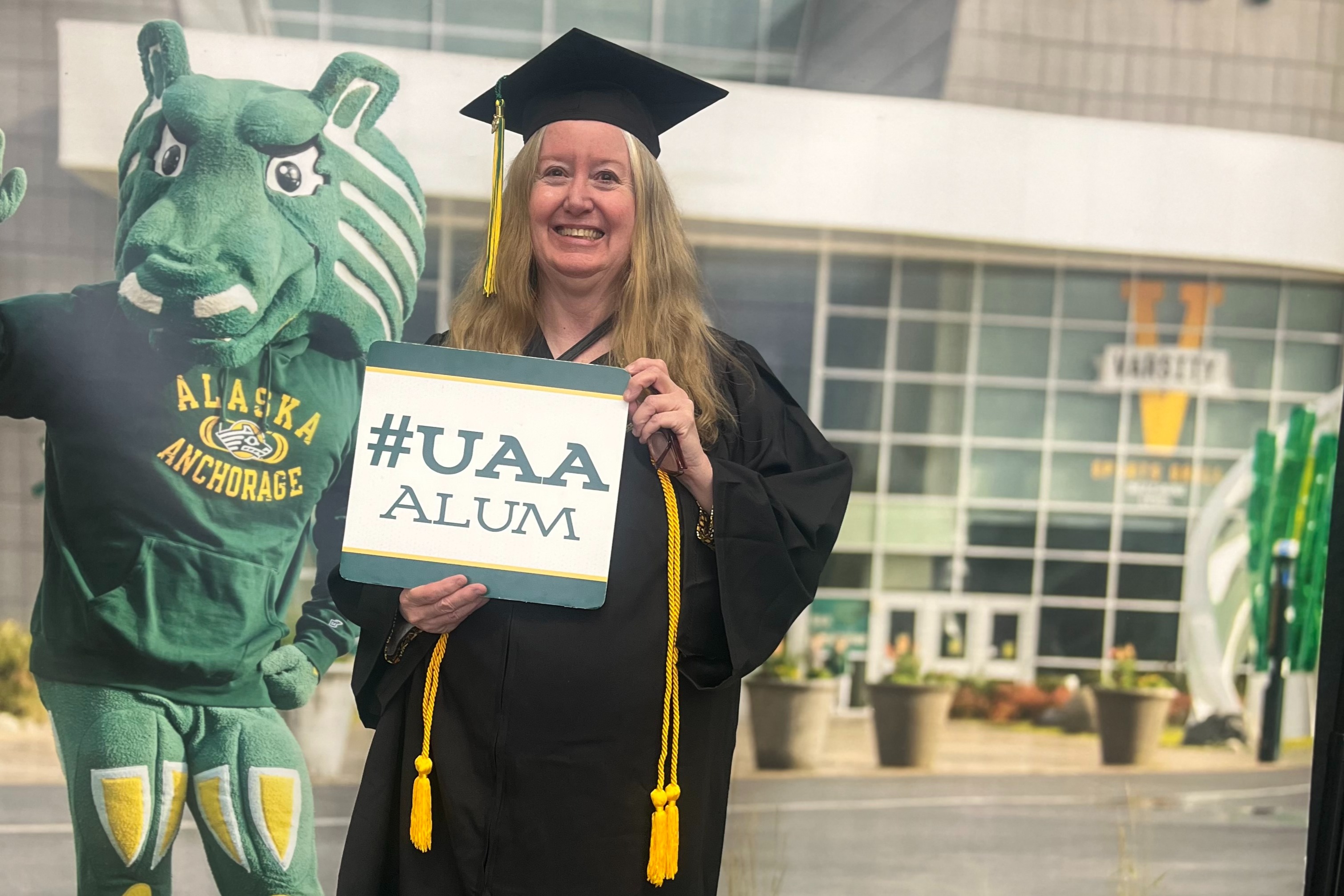 Billie stands in front of a UAA backdrop featuring Spirit the Seawolf. She is in full graduation regalia and holding a sign that reads "#UAA Alum."