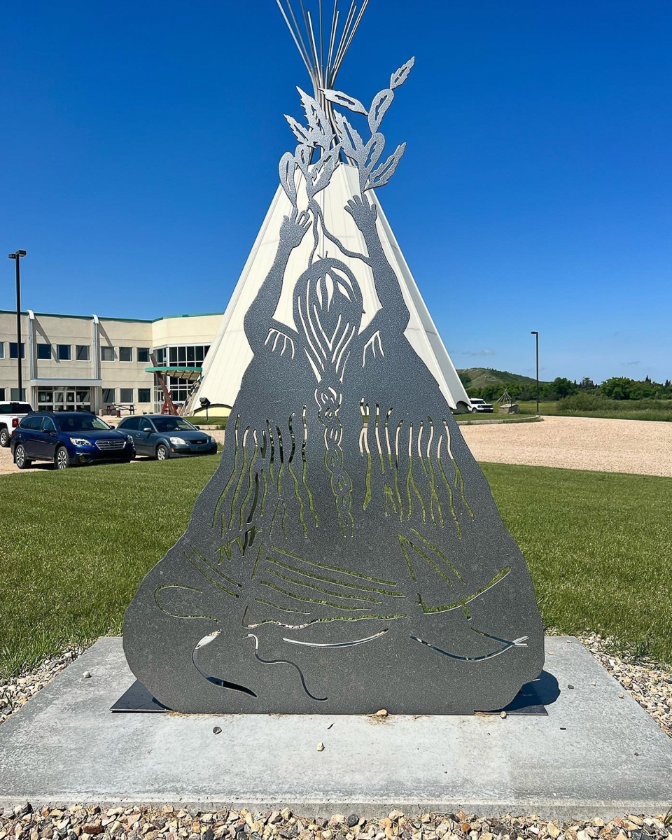 metal statue of indiginous woman throwing feathers in air in front of teepee in front of building