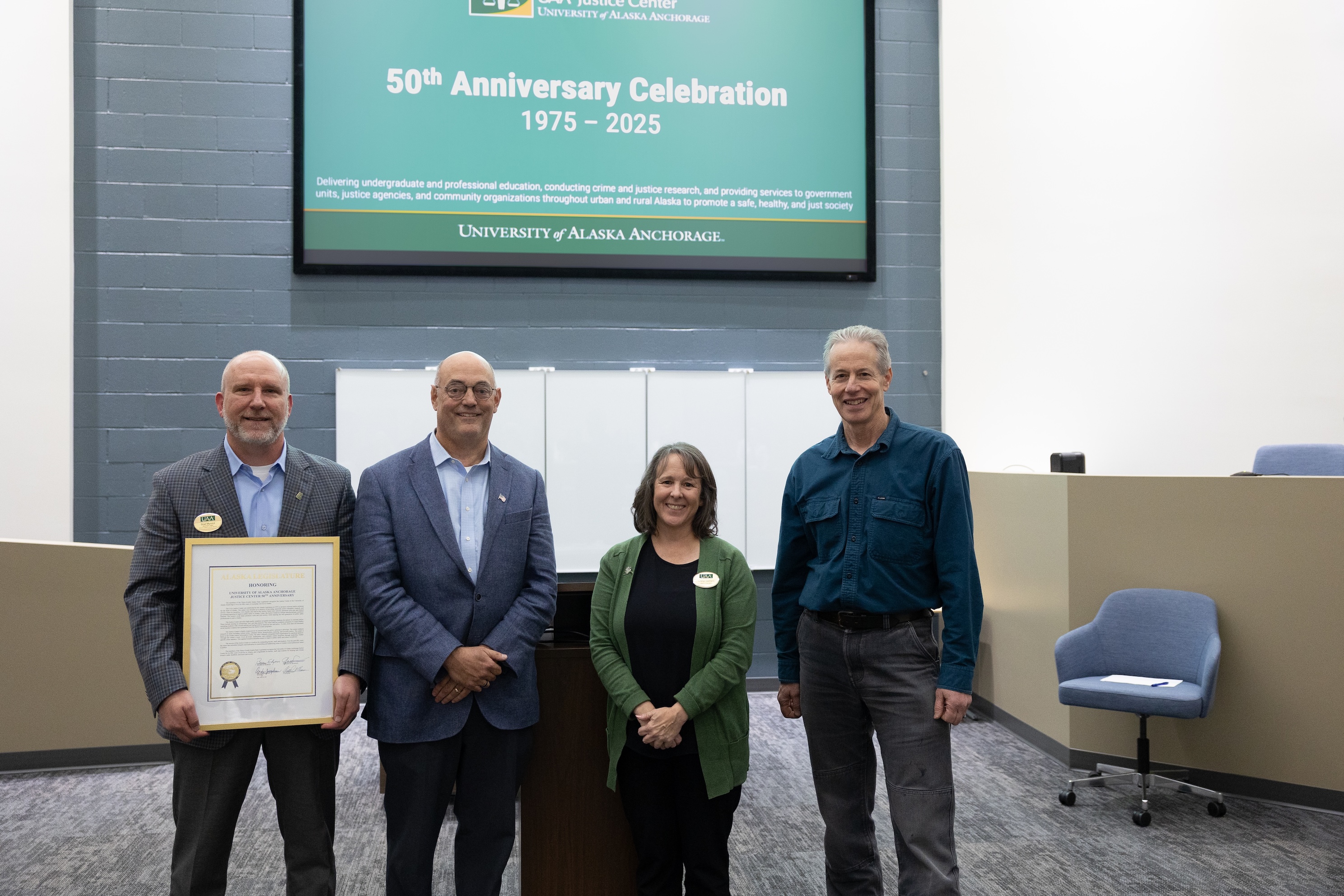 Assistant Dean Brad Myrstol, Representative Andy Josephson, Chancellor Cheryl Siemers, and Senator Matt Claman stand side by side in front of a podium in the Havelock Simulation courtroom. Myrstol is holding the framed legislative citation.