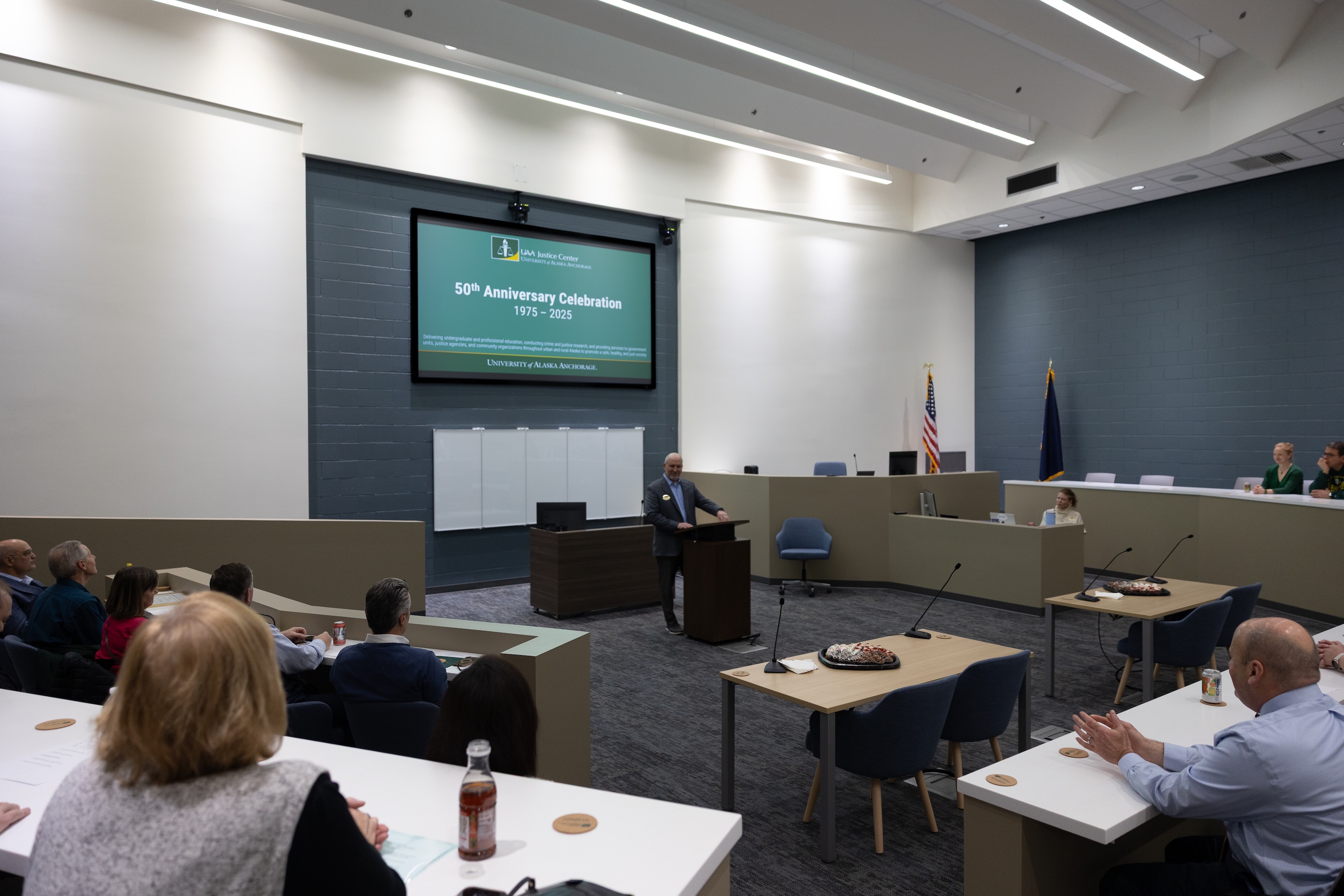 Assistant Dean Brad Myrstol stands behind a podium delivering opening remarks at the legislative citaiton event. Before him are audience members seated at desks around the room.