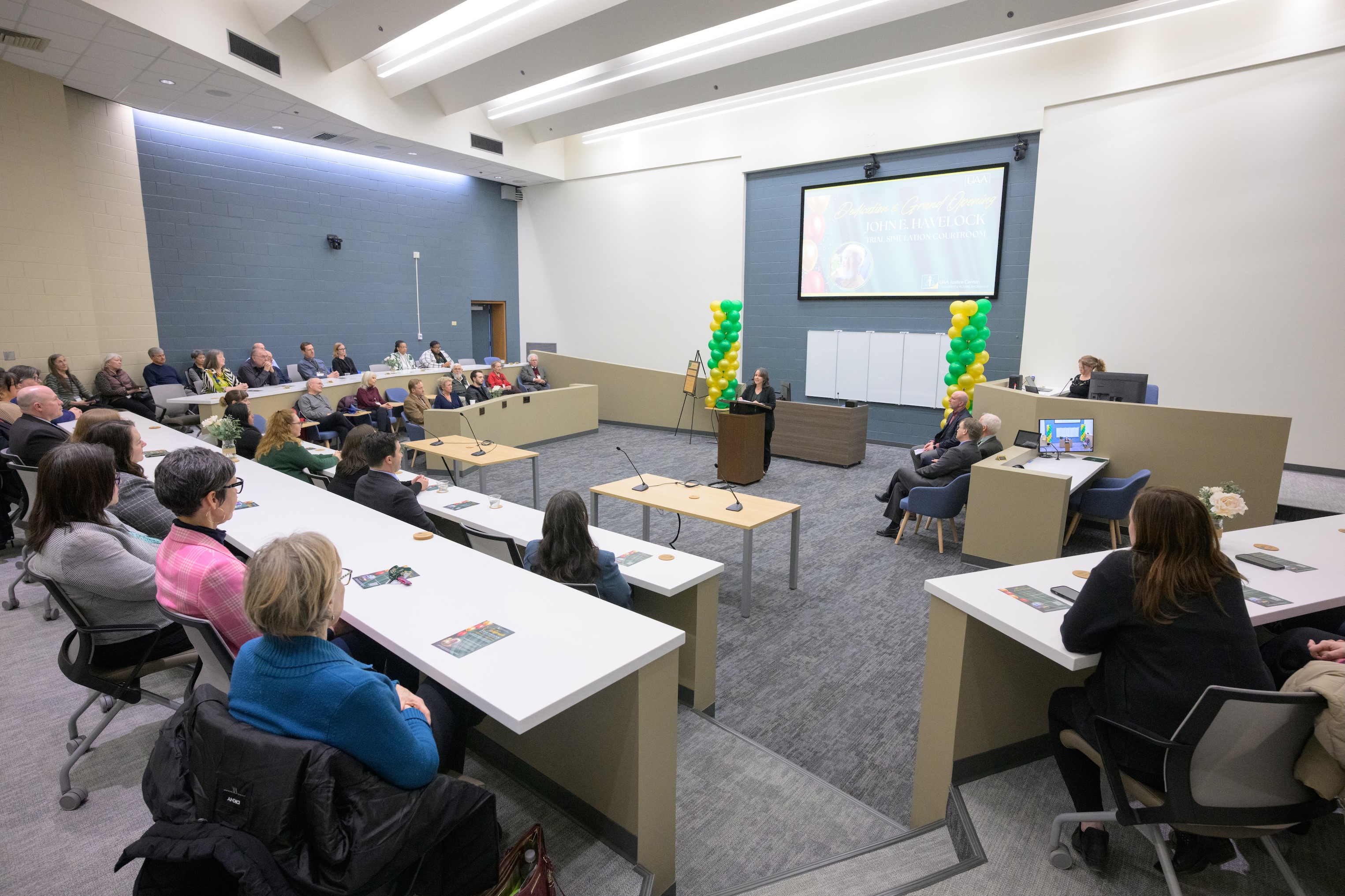 Interim Chancellor Cheryl Siemers stands at the podium in the Havelock Courtroom and addresses an audience of faculty, staff, and others at the dedication event. Behind her are celebratory balloon pillars. 