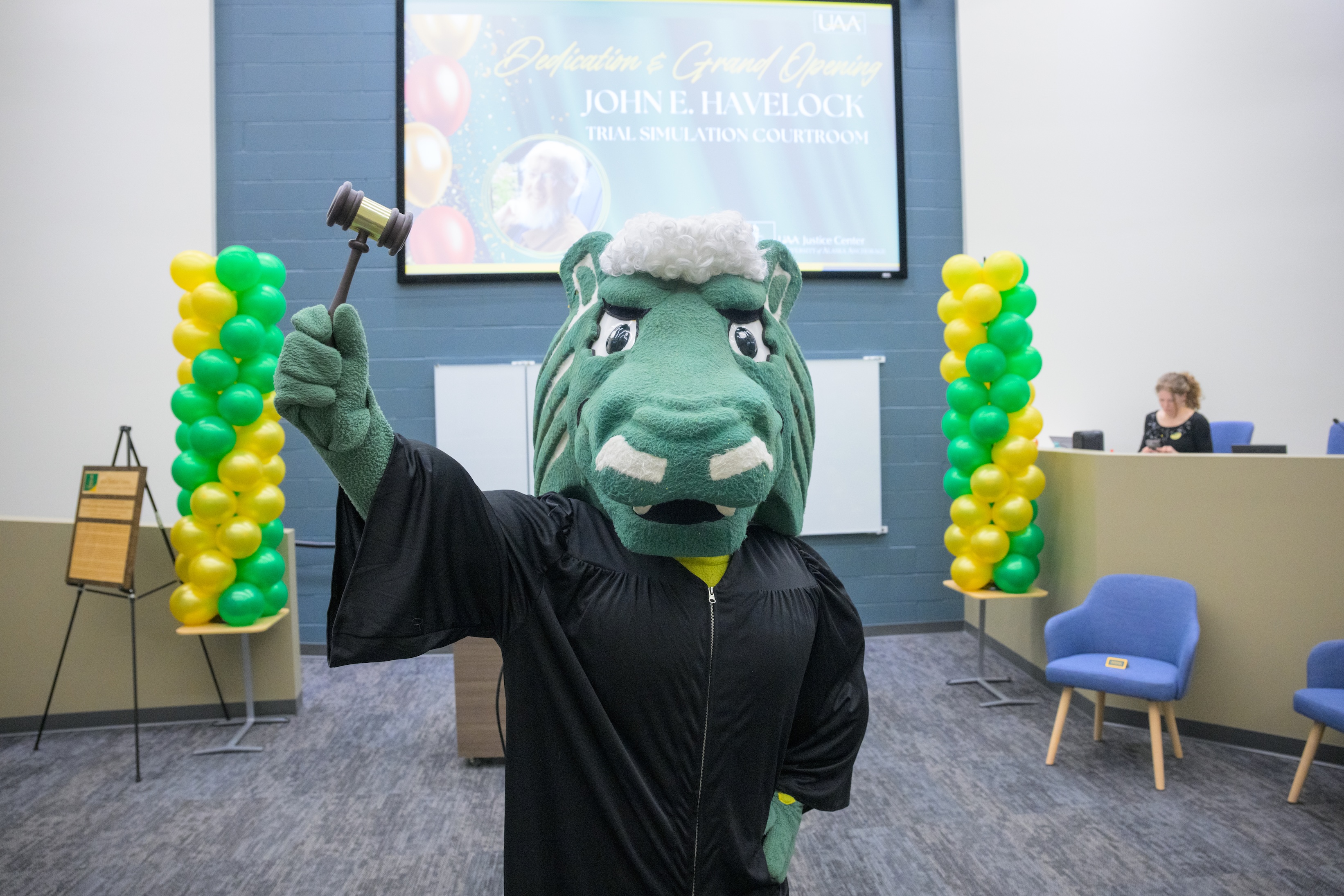 A person dressed as Spirit the Seawolf stands in the Havelock classroom wearing black robes and holding a gavel. Behind them are celebratory balloon pillars and a slide about the dedication and grand opening. 