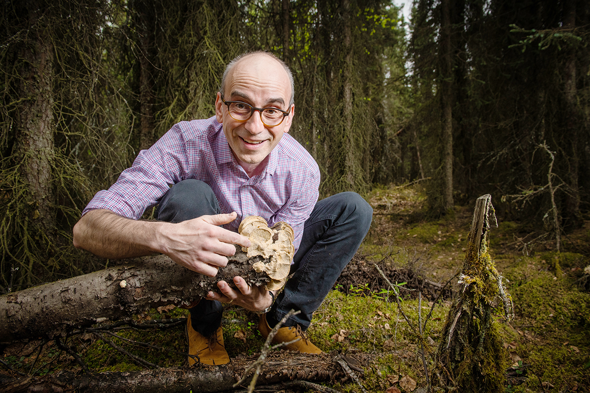Philippe Amstislavski crouches in the forest, smiling. He is propping up one end of a fallen tree with mushrooms growing on it, and pointing excitedly to the mushrooms. 
