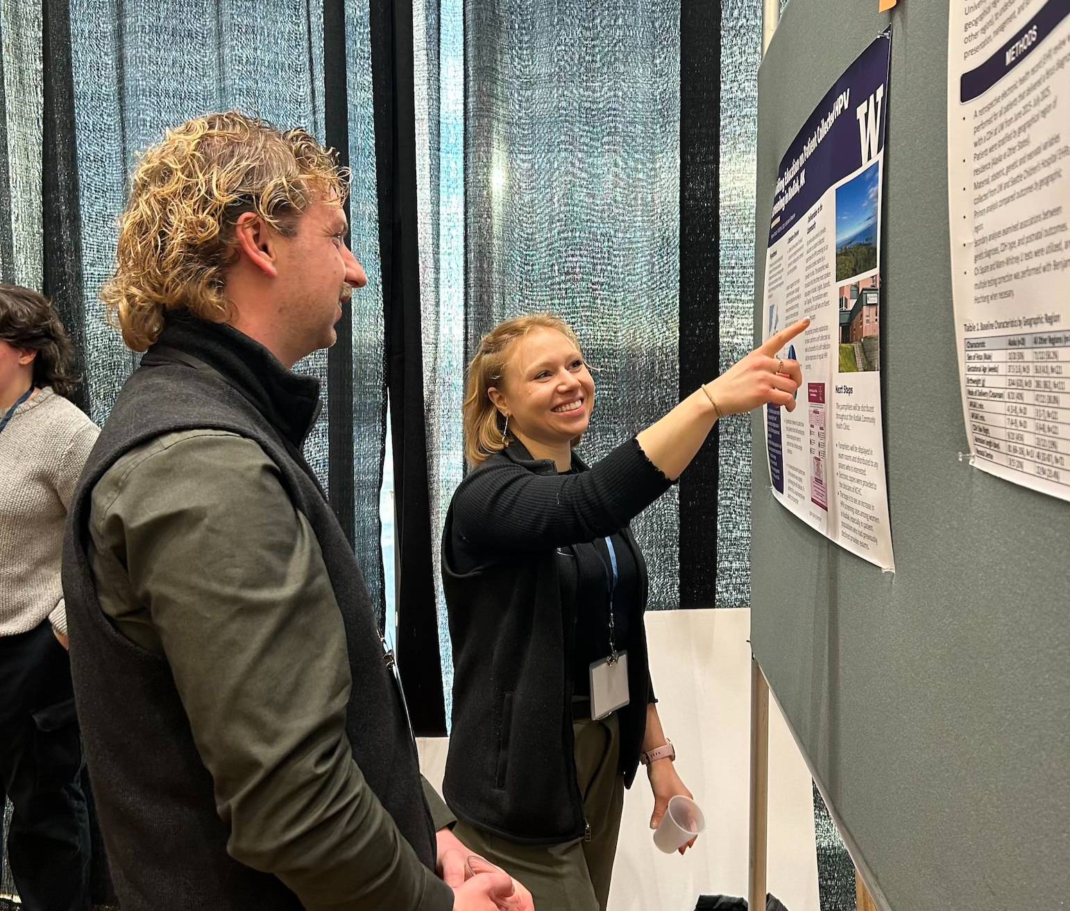 A student stands in front of her poster and points to it. Next to her, another student stands and looks at the poster with his hands folded. 