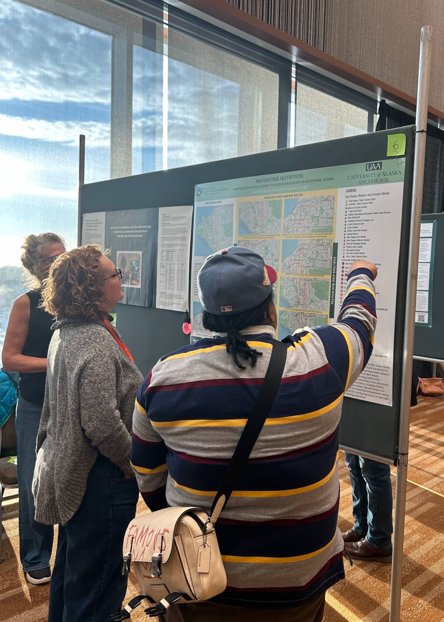 A UAA student stands in front with her back to the camera. She is pointing to her poster. A conference attendee stands next to her and looks at her poster. 