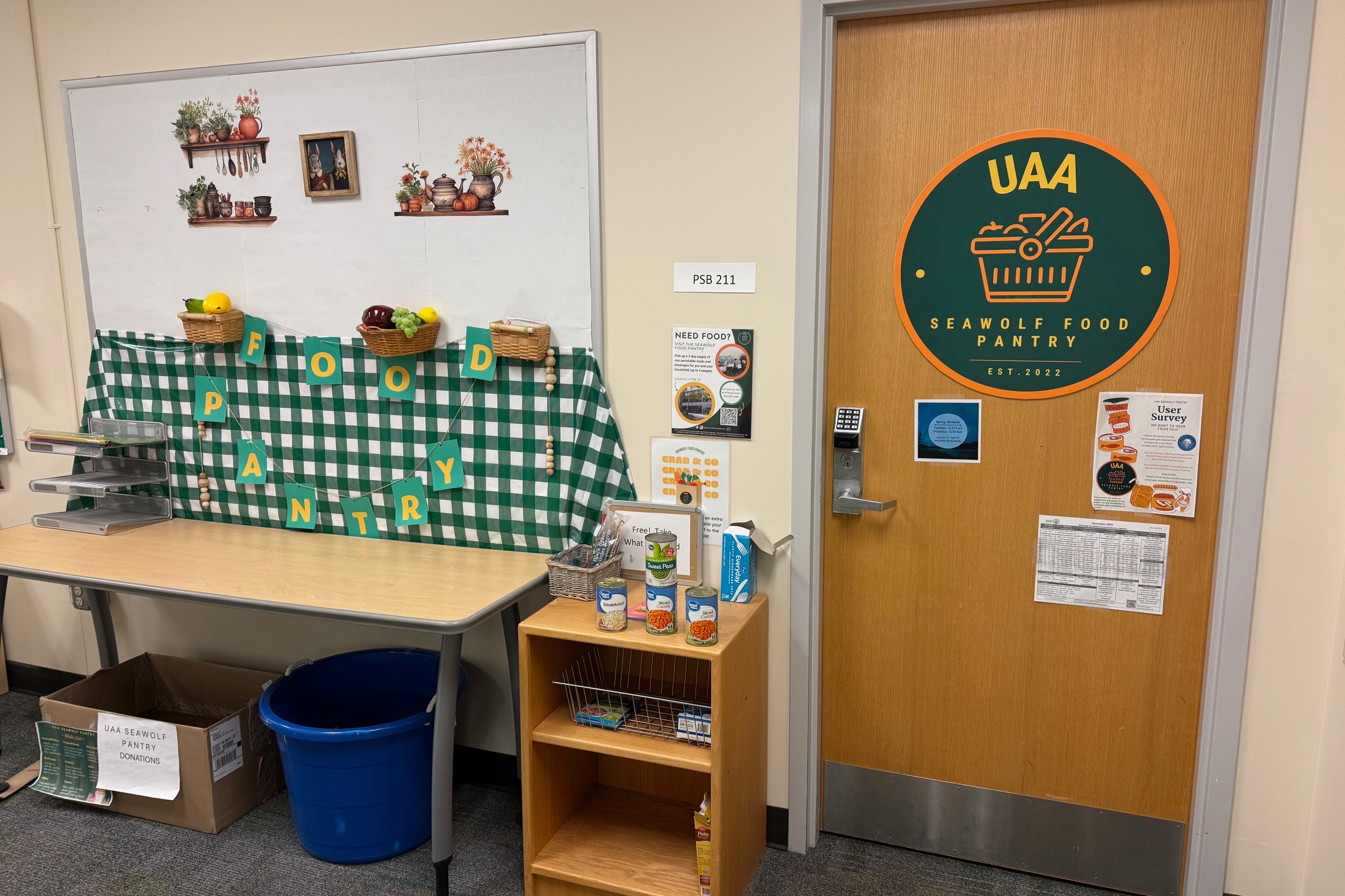 Photo shows a closed door with a sign on it that reads "UAA Seawolf Food Pantry." A small table next to the door has several canned food items and decorations. 
