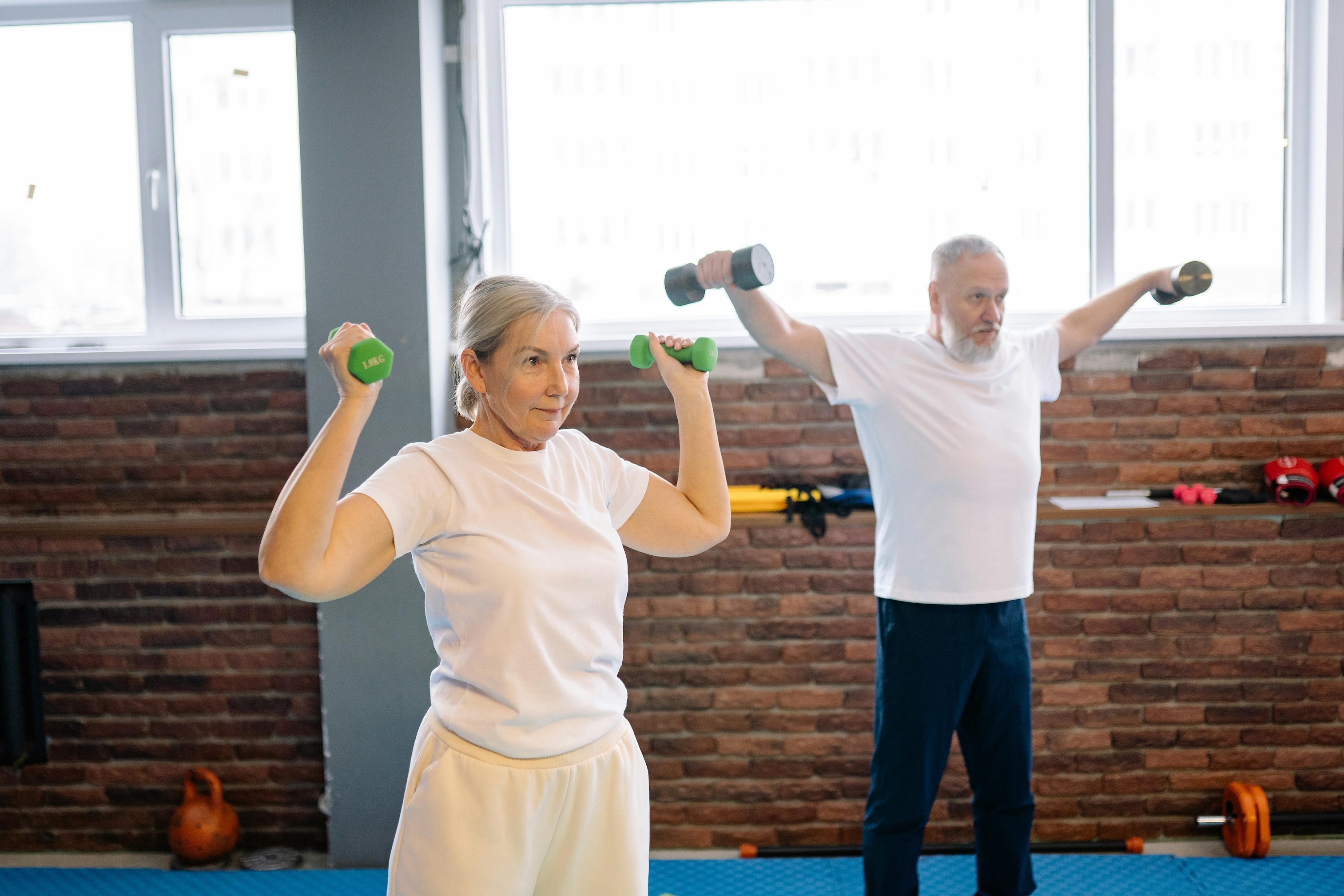 An older man and woman are pictured standing in a gym. Both are holding small dumbbells and wearing workout clothes. 