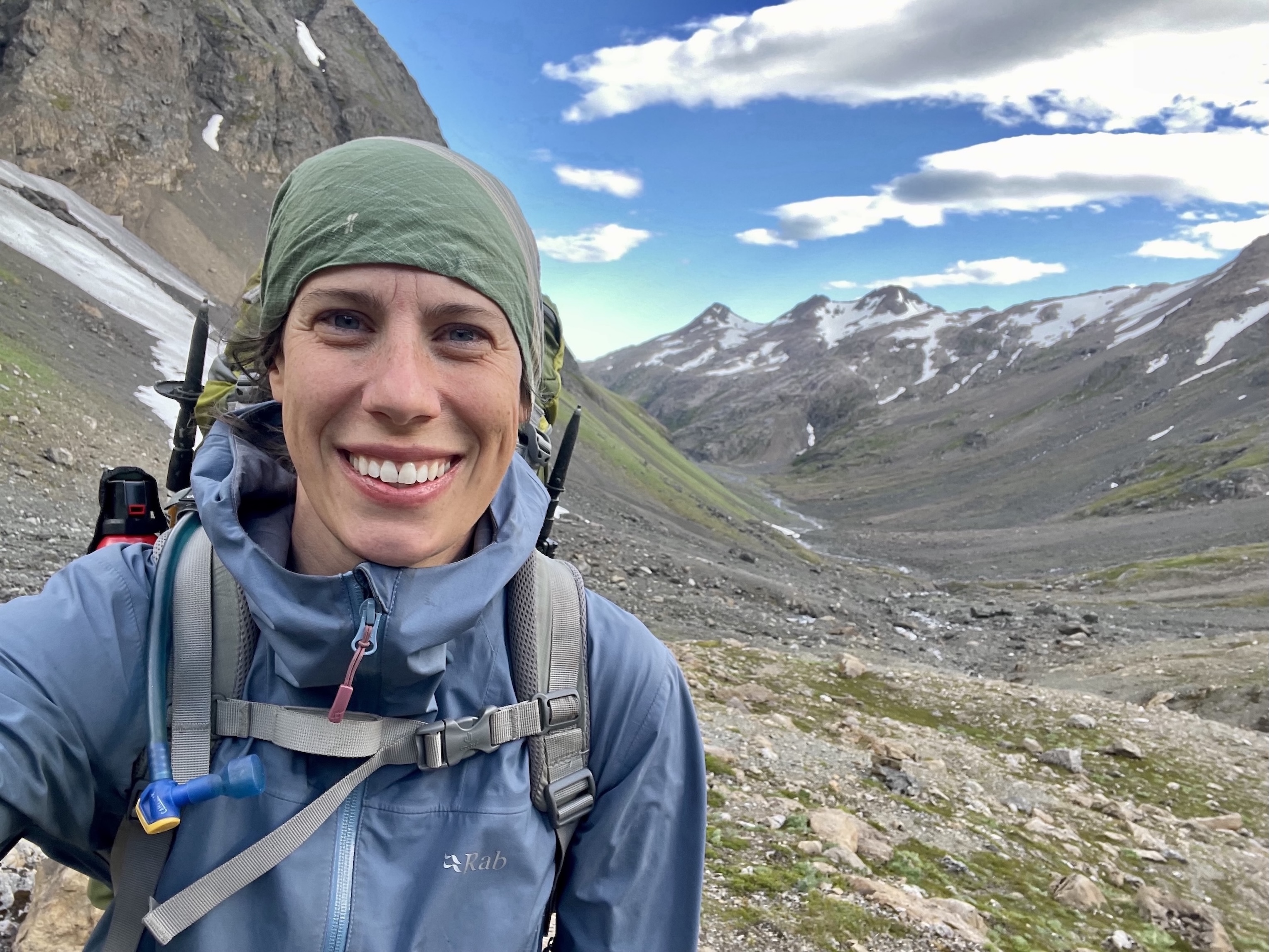 Tara smiles at the camera while she takes a selife. She is wearing a backpack and outdoor gear, and standing in a pass with mountains on either side. 