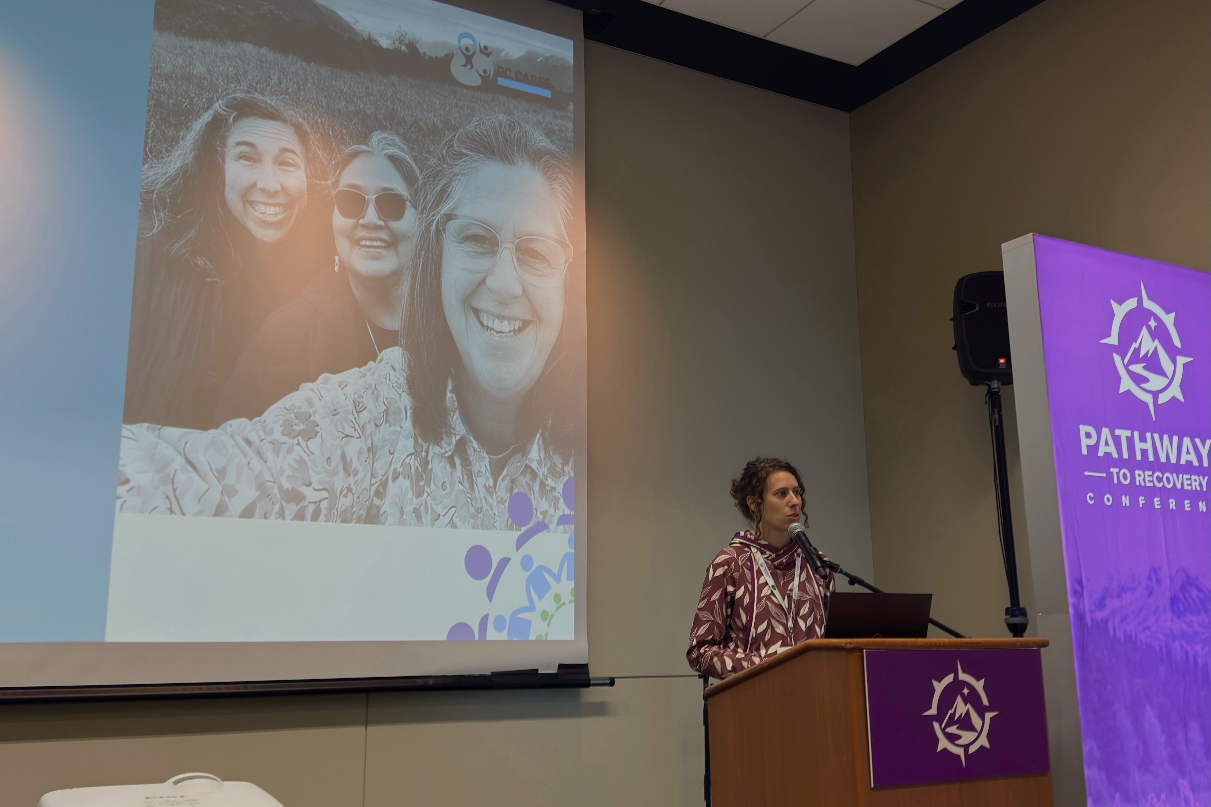 Tara stands behind a podium and speaks into a microphone at the Pathways to Recovery Conference. Behind her is a slide deck showing three smiling women. 