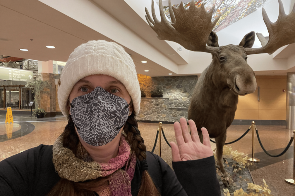 Monica Roe stands in the Ted Stevens International Airport in Anchorage. She is dressed warmly, with a scarf and beanie hat, and is wearing a mask. She stands in front of a taxidermied moose and raises a hand. 