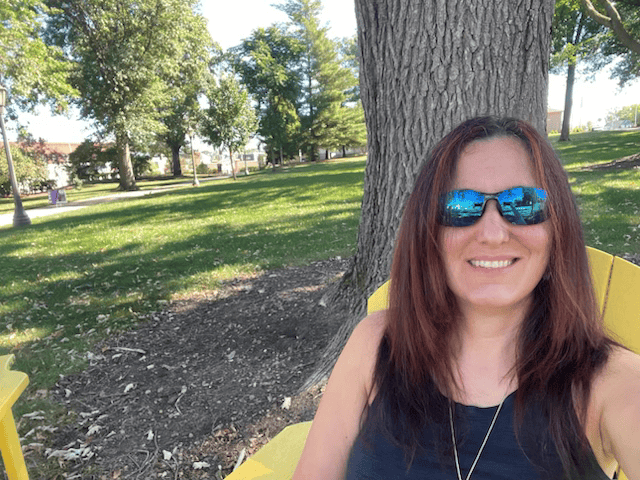 Monica Roe sits in a yellow lawn chair in an open outdoor space in front of a tree. She is wearing a tank top and sunglasses and smiling. 