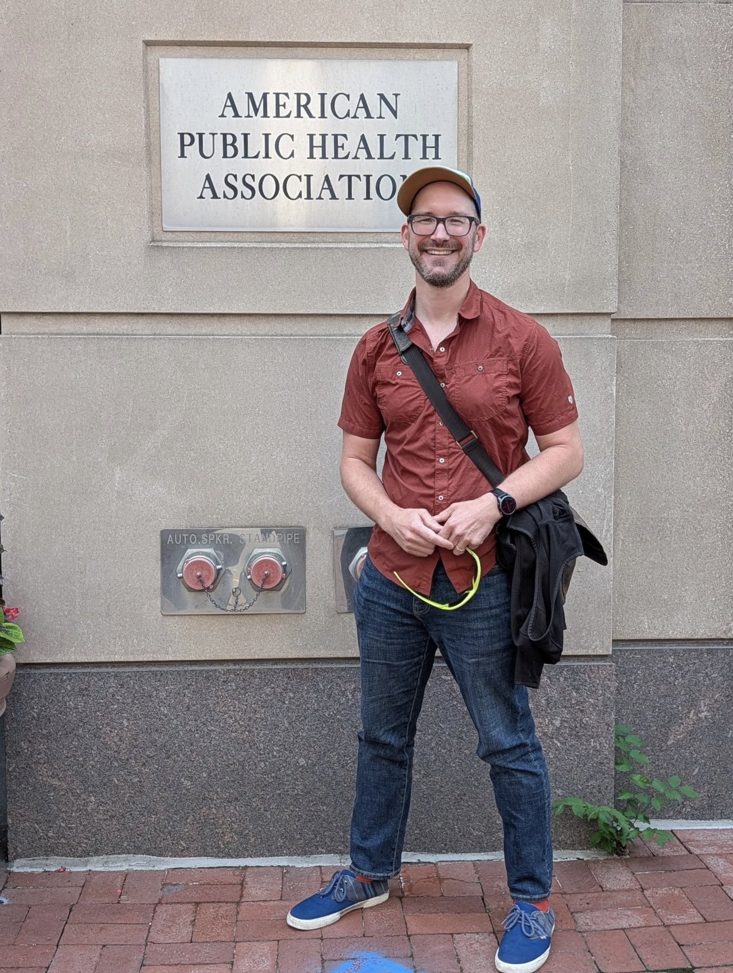 A man in blue jeans, red button up, blue sneakers, a baseball cap, and glasses poses in front of a building with a sign "American Public Health Association"