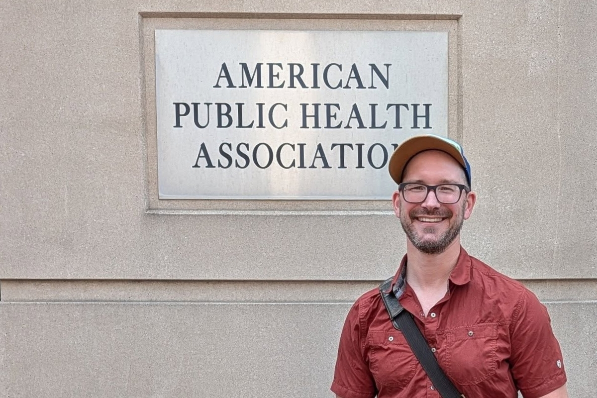 A man in a red button down, baseball cap, and glasses posing in front of a building with a sign "American Public Health Association"