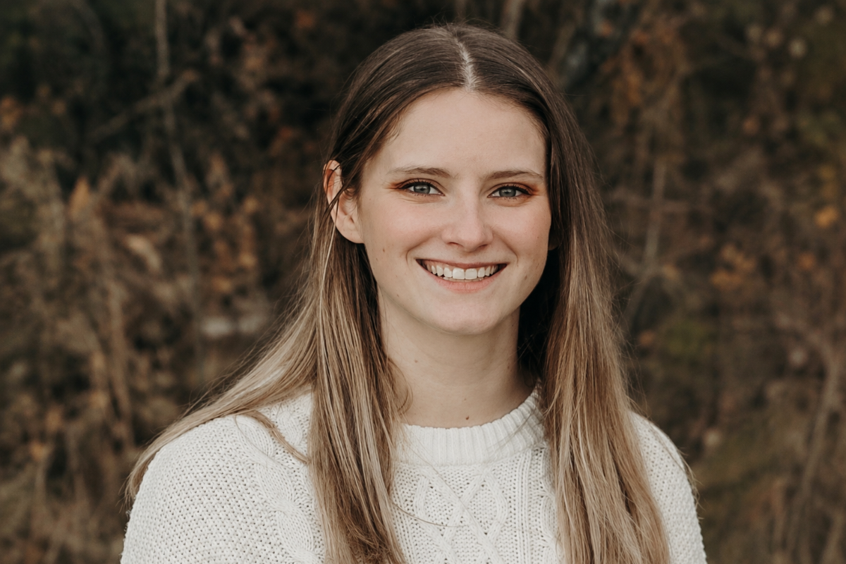 A woman with long brown hair in a white sweater smiles in front of an autumn background