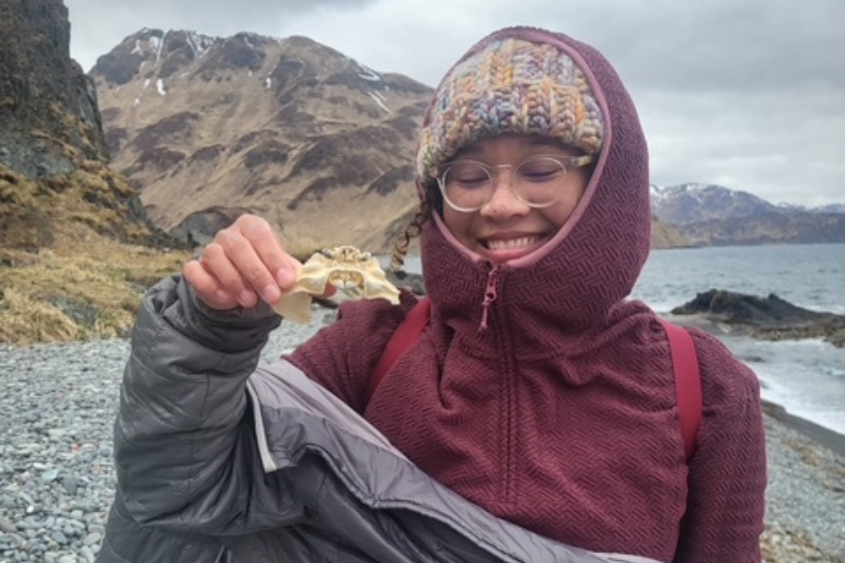 A woman in a purple jacket, knit hat, and glasses smiles on a rocky beach holding a shell