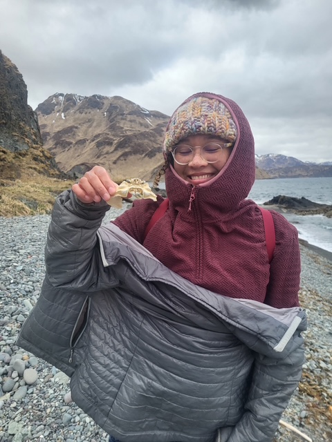 A woman in a purple jacket, knit hat, and glasses smiles on a rocky beach holding a shell