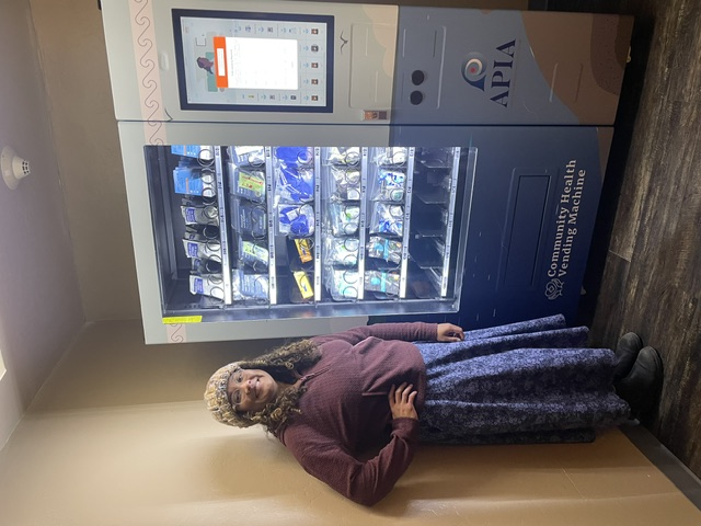 A woman in a purple jacket, long skirt, and knit hat poses in front of a lit vending machine