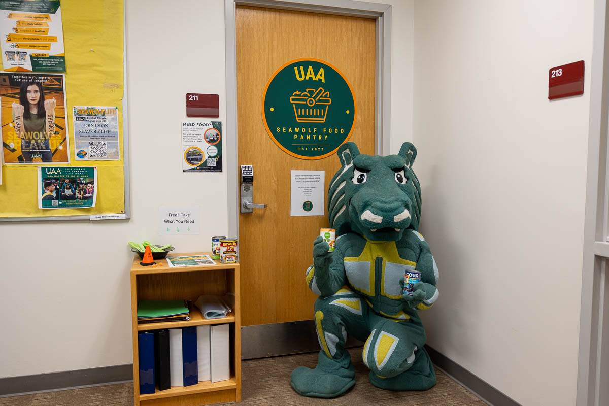A person in a Spirit the Seawolf costume kneels in front of the food pantry door holding a can of beans in each hand. 