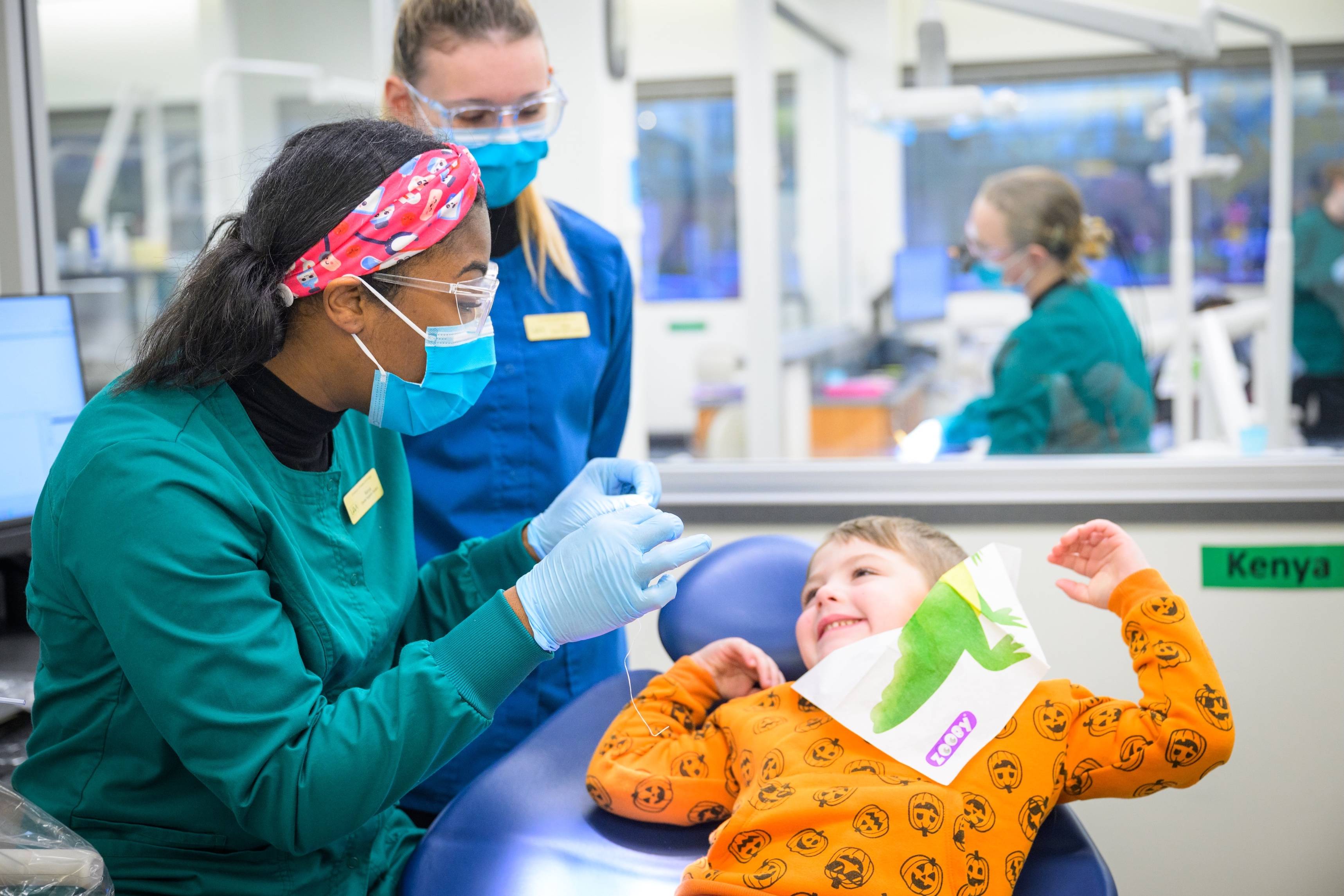 A dental hygiene student wearing a green long-sleeved scrub top, surgical mask, headband, and gloves prepares to floss a young child wearing a Halloween shirt and dental bib with a dinosaur on it. 