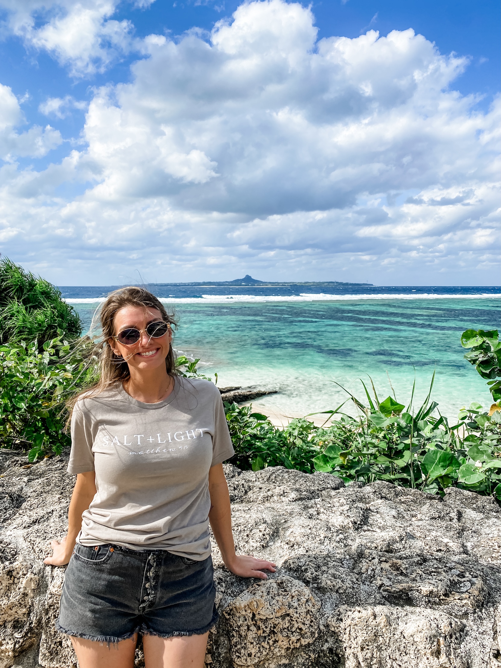 Ambra McMillian leans against a boulder. Behind her is a tropical beach, lush foliage, and a blue sky with clouds. 