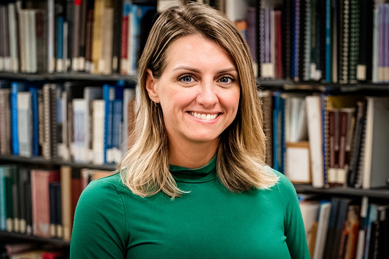 Ambra McMillian smiles at the camera in front of shelves filled with books an journals. She has shoulder length blonde hair and is wearing a green sweater. 