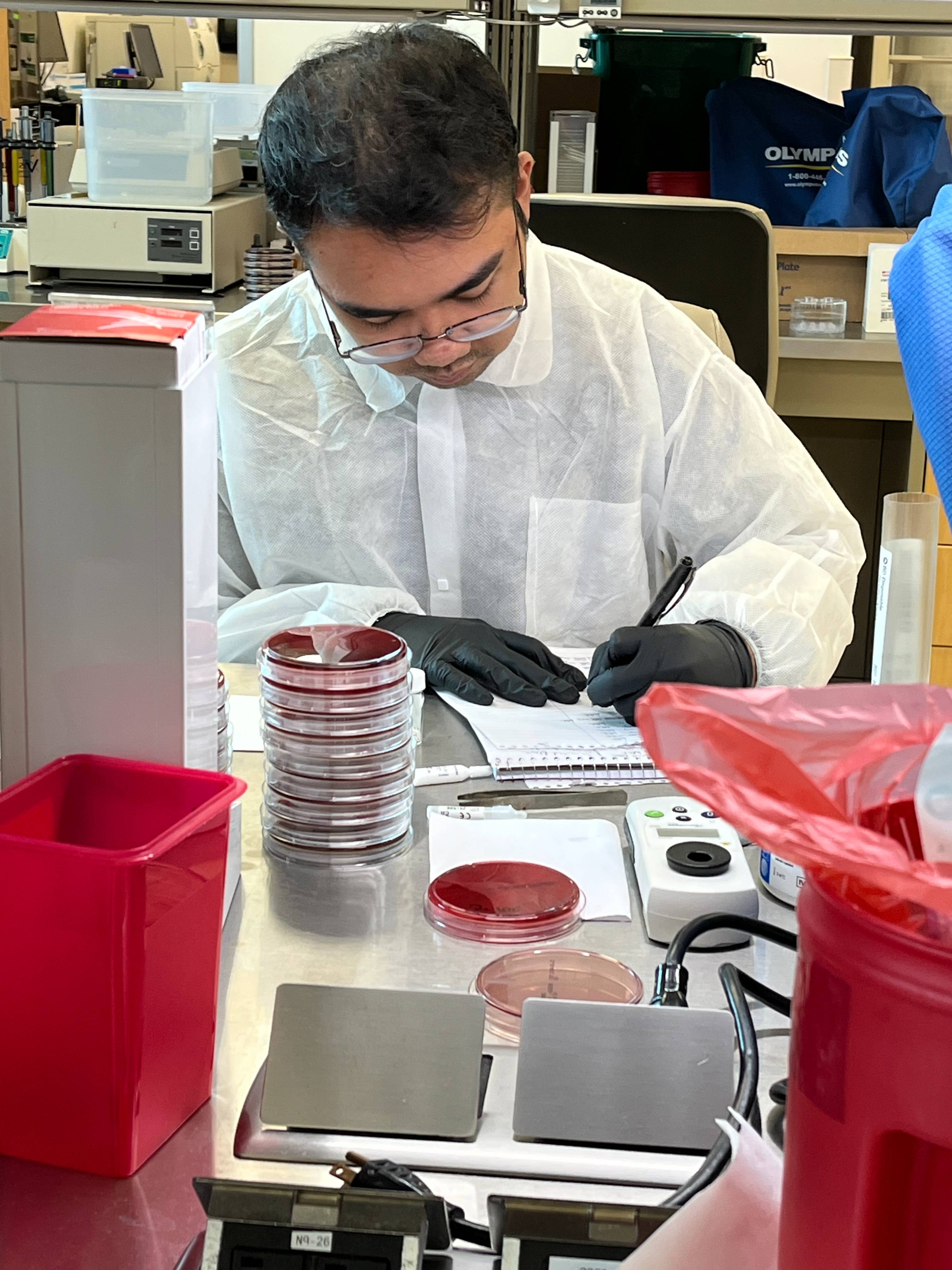 Medical Laboratory Science student Rieluis Martin sits at a lab bench and makes notes in a notebook. He is wearing a white sterile gown and lab gloves. In front of him is a stack of petri dishes and instruments.