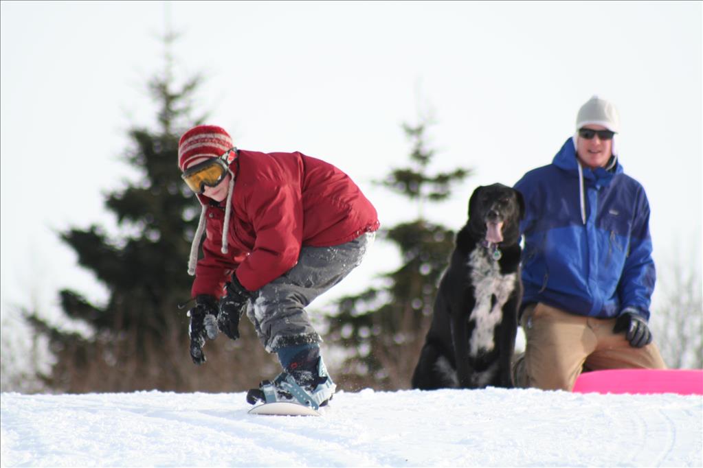 Volunteer and dog watching child snowboard in snow 
