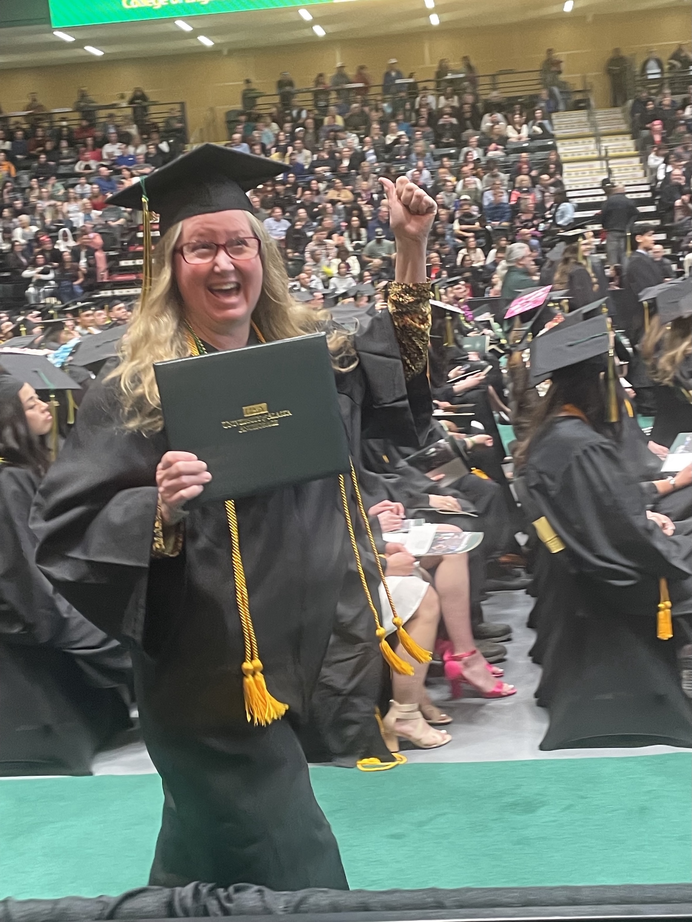 Billie walks down the aisle at commencement in full graduation regalia. She is holding her diploma in one hand and doing a fist pump with the other while smiling broadly.