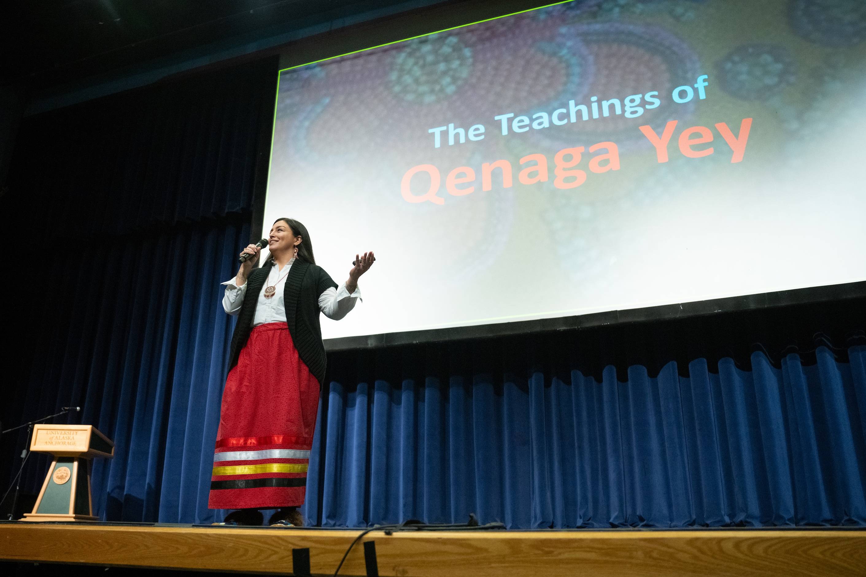 Jessica Ross stands on a stage. She is holding a microphone and wearing traditional Indigenous clothing. Behind her is a projector screen that reads "The Teachings of Qengaga Yey."