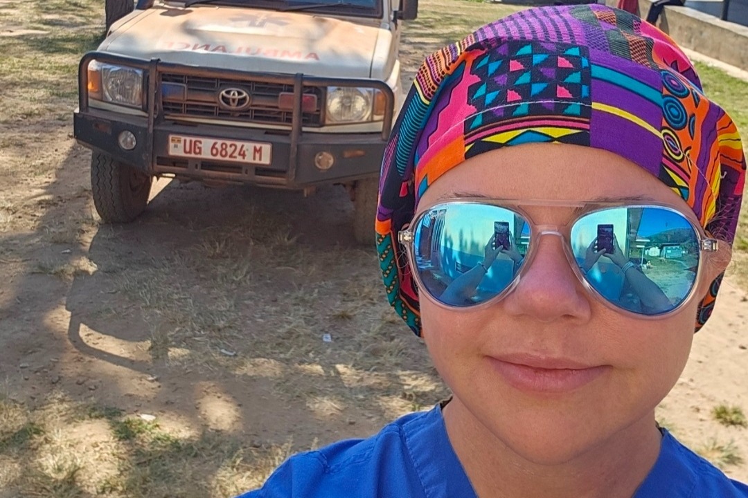 Kristen Alcorn is wearing aviator sunglasses, a colorful head scarf, and blue scrubs. She is standing outside in front of a dusty white SUV labeled "Ambulance."