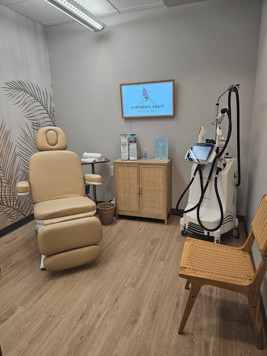 Photo depicts a room with wood floors and neutral gray walls. On one side is a padded patient chair with arm and headrests. On the back wall is a screen that reads "Northen Grace Medical Spa." In the corner across from the chair is a standing electronic medical device.