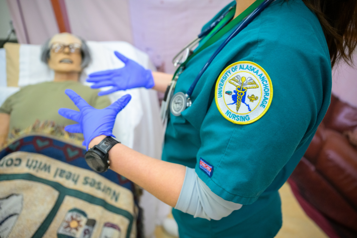 A person in green "University of Alaska Anchorage nursing" scrubs with latex gloves on speaking in front of a manikin