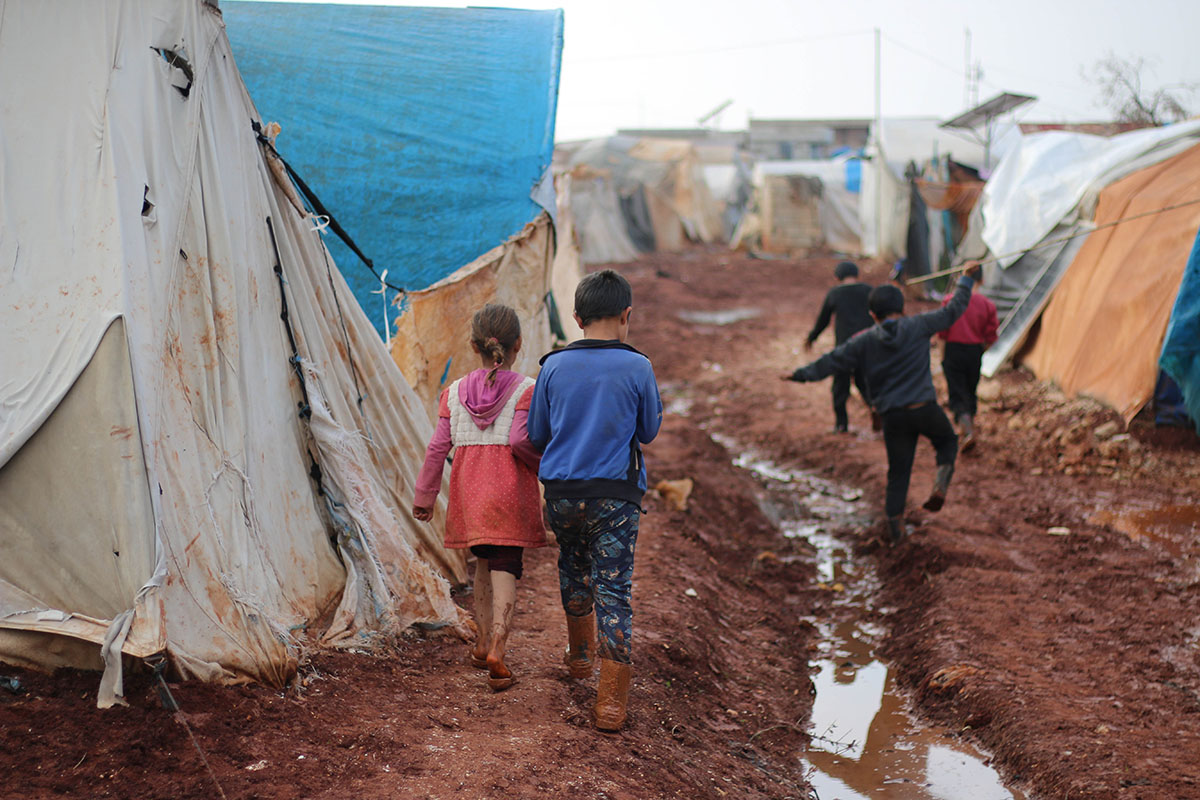 Children walking on a muddy pathway with tents