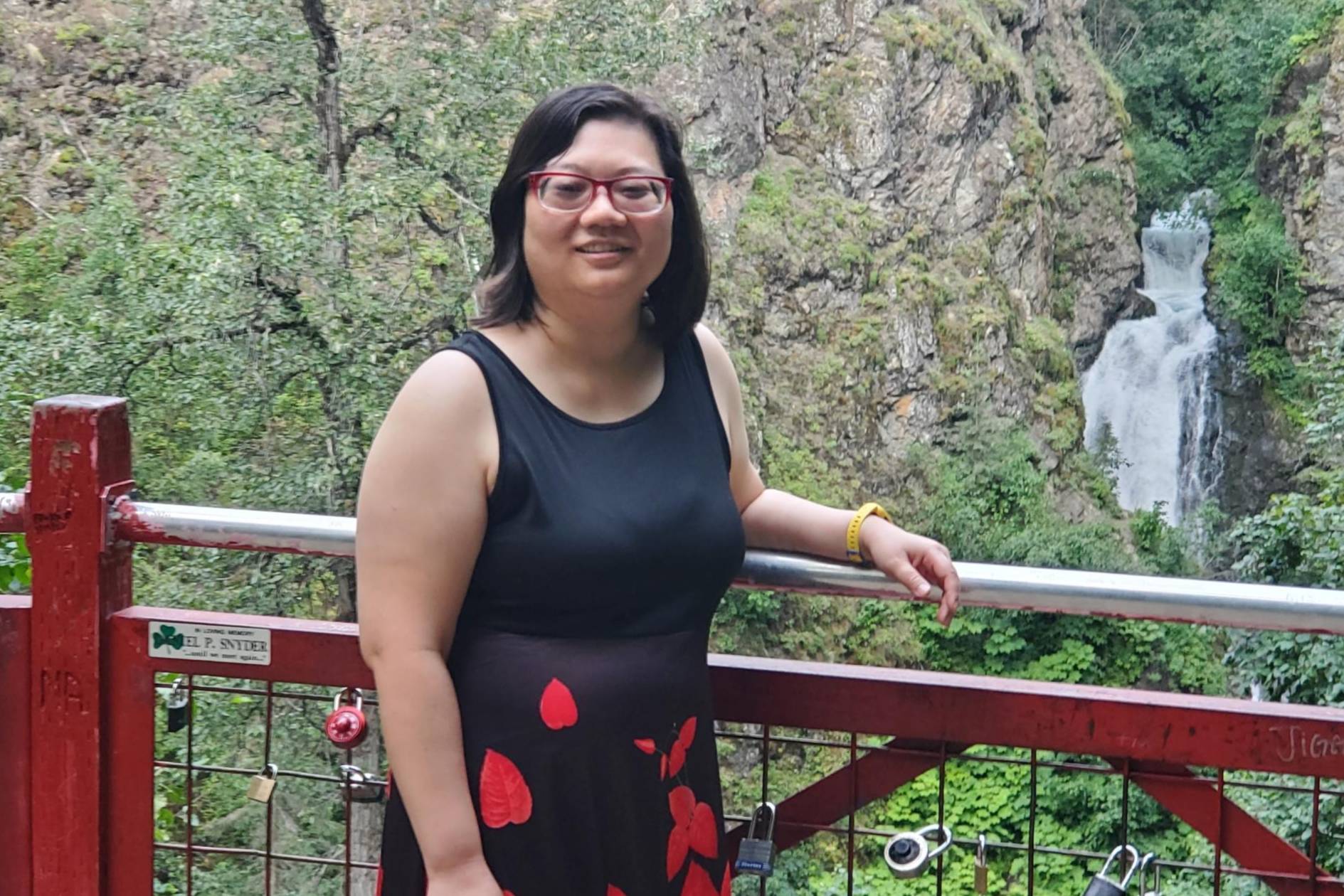 Master of Social Work student MiLee Yu stands on a red bridge with one arm on the railing. In the distance behind the bridge is a waterfall and vegetation on a cliff. 