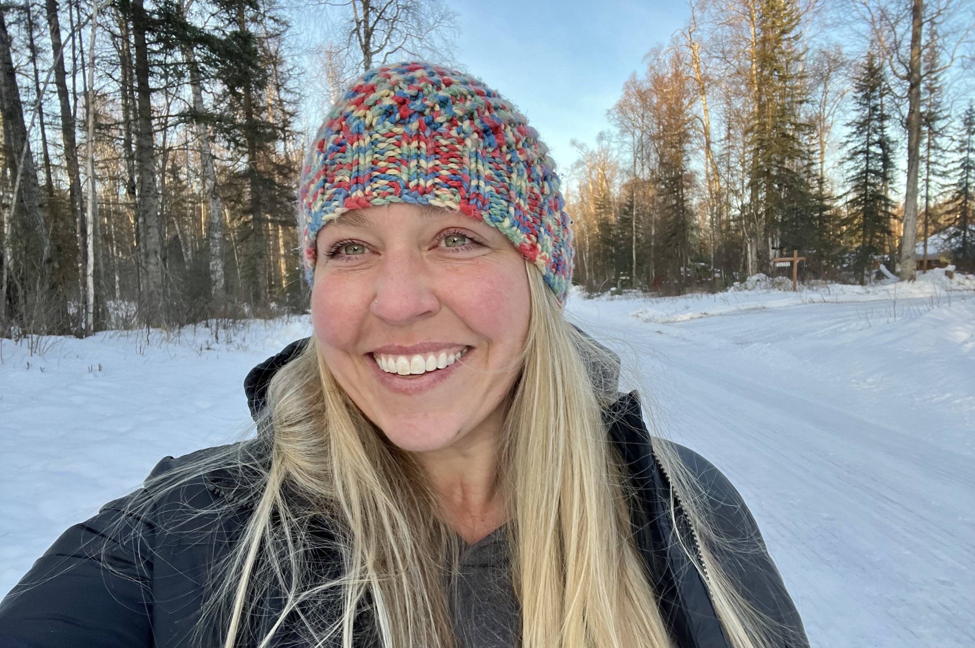 Dacia Davis stands on a snowy road in Talkeetna with trees on either side. She is wearing a multicolored knit beanie hat and a black coat and is smiling.