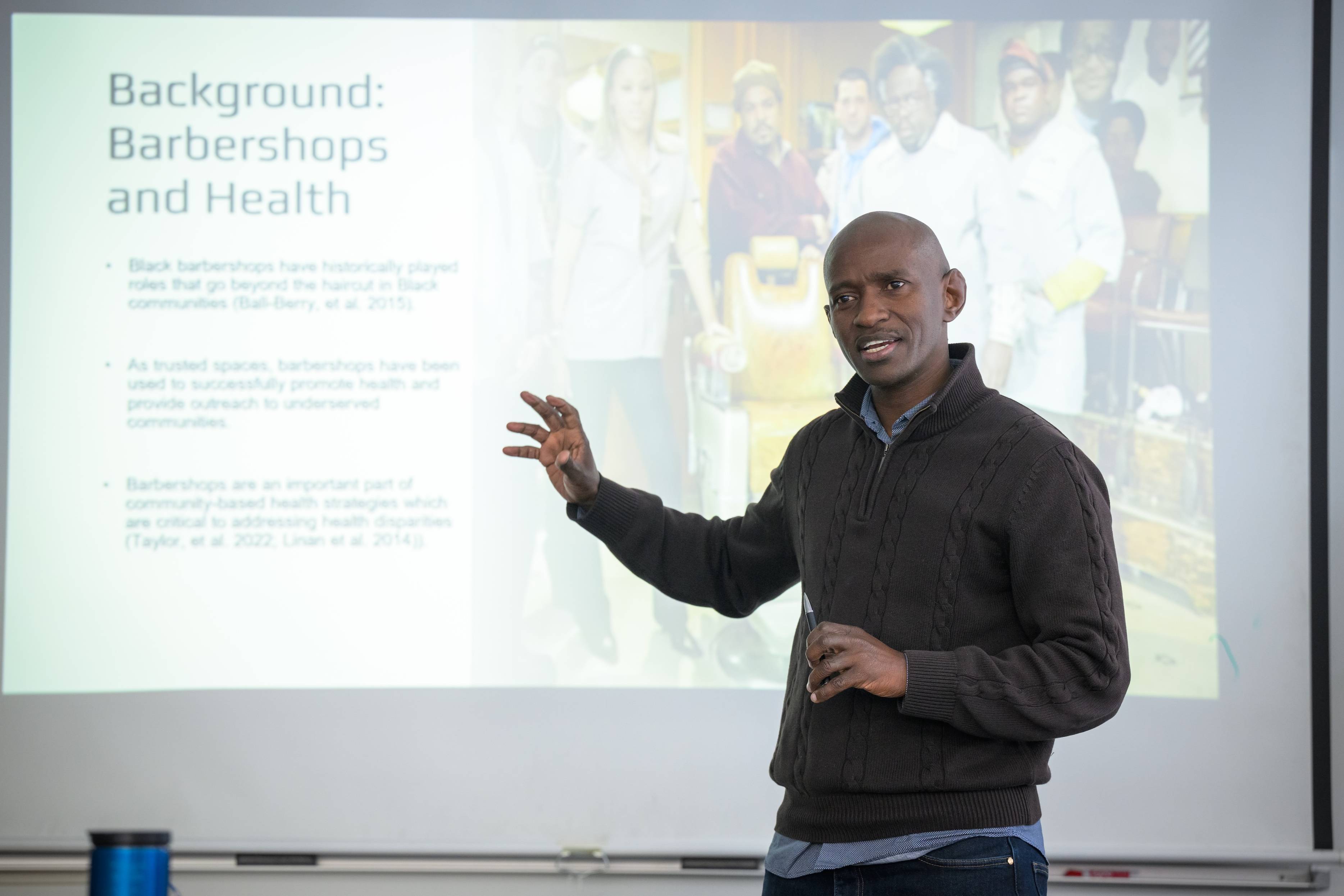 Amana Mbise stands in front of a screen with a slide that reads "Background: Barbershops and Health." He is gesturing and speaking to an audience (not pictured)