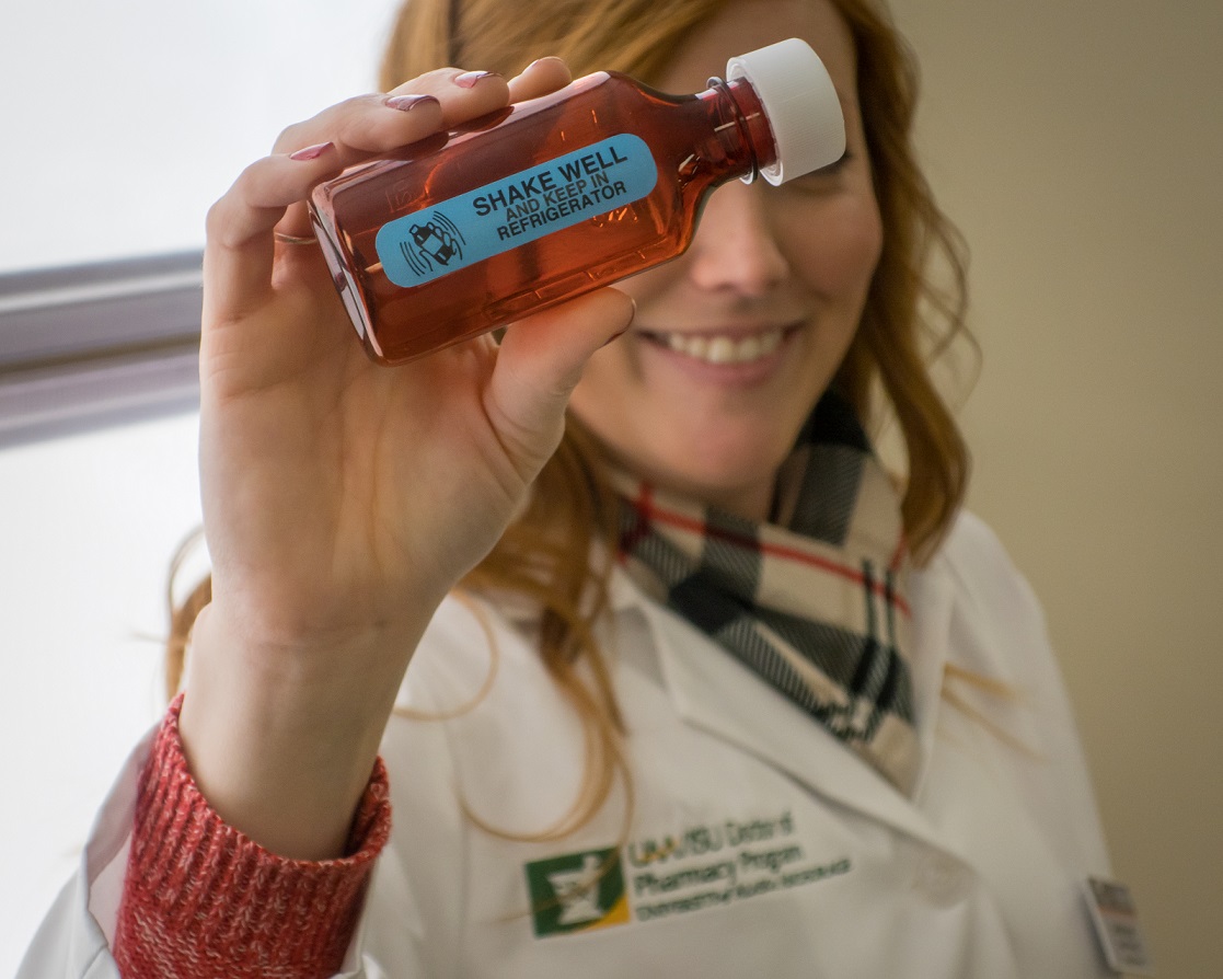 A pharmacy student in a lab coat holds a bottle labeled "shake well"