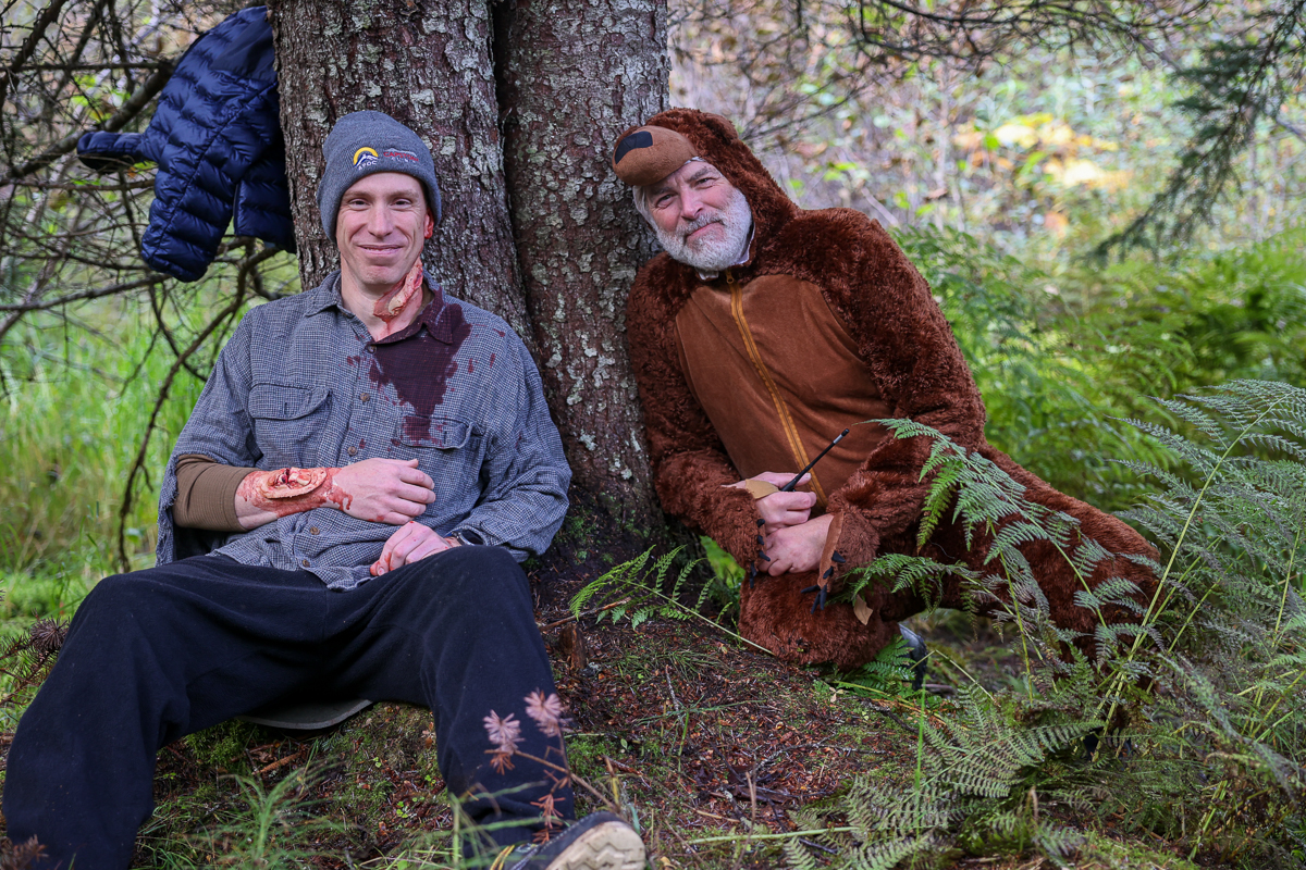 a man with fake injuries and a man in a bear costume lean against a tree