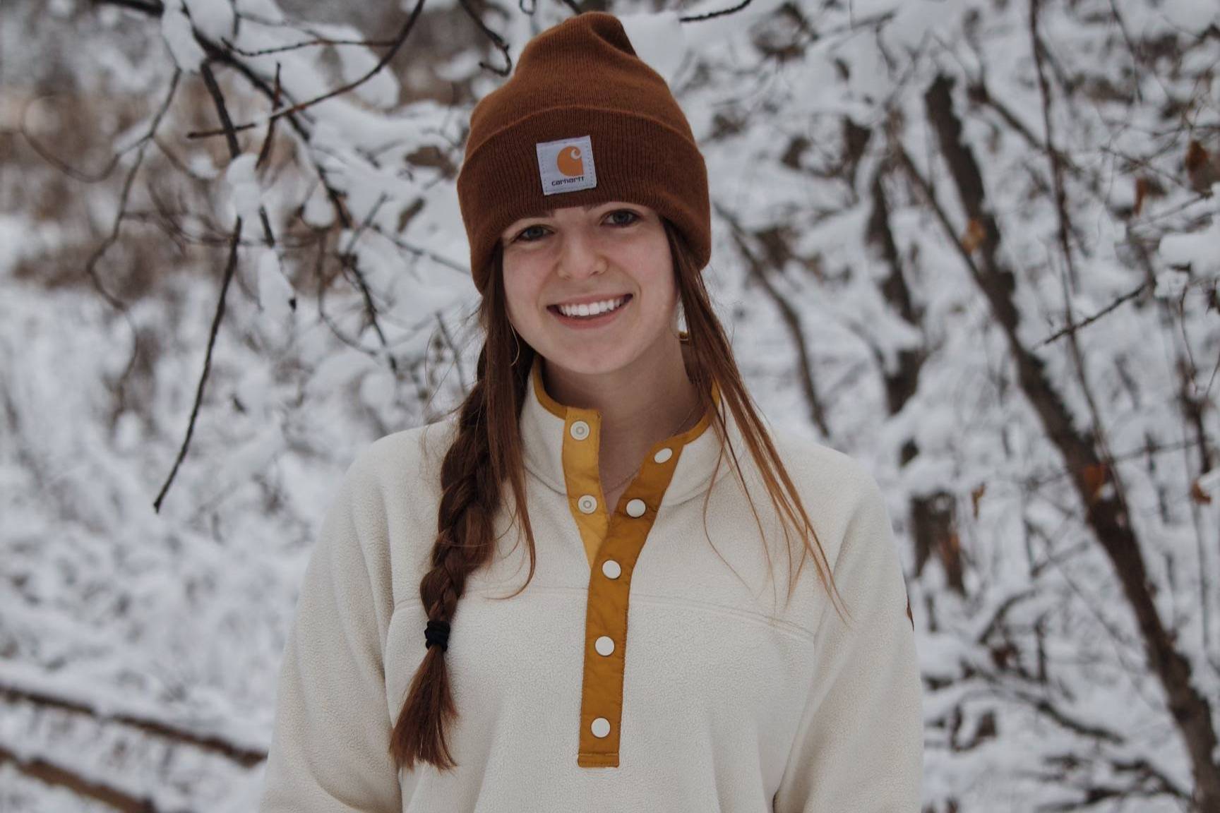 Abigail Crawford wears a white sweater and Carhartt beanie. She is standing in front of a snowy wooded background and smiling at the camera. 