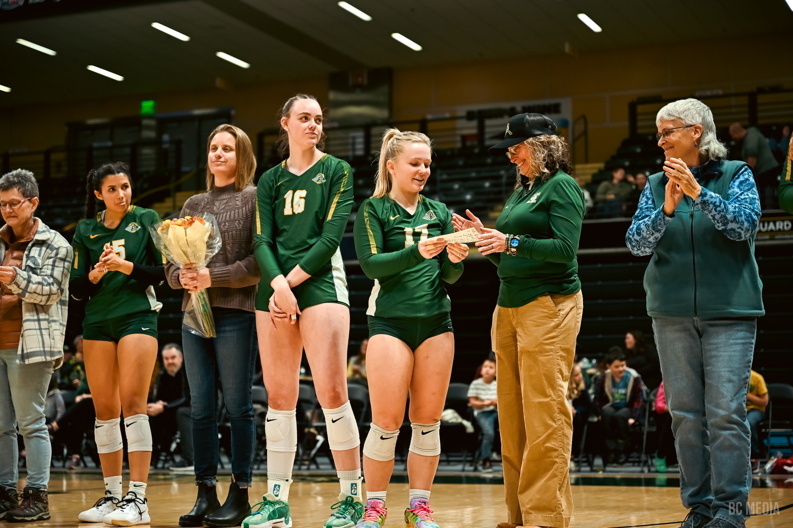 UAA volleyball athletes in green jerseys stand next to faculty. One player presents a piece of paper to a faculty member.