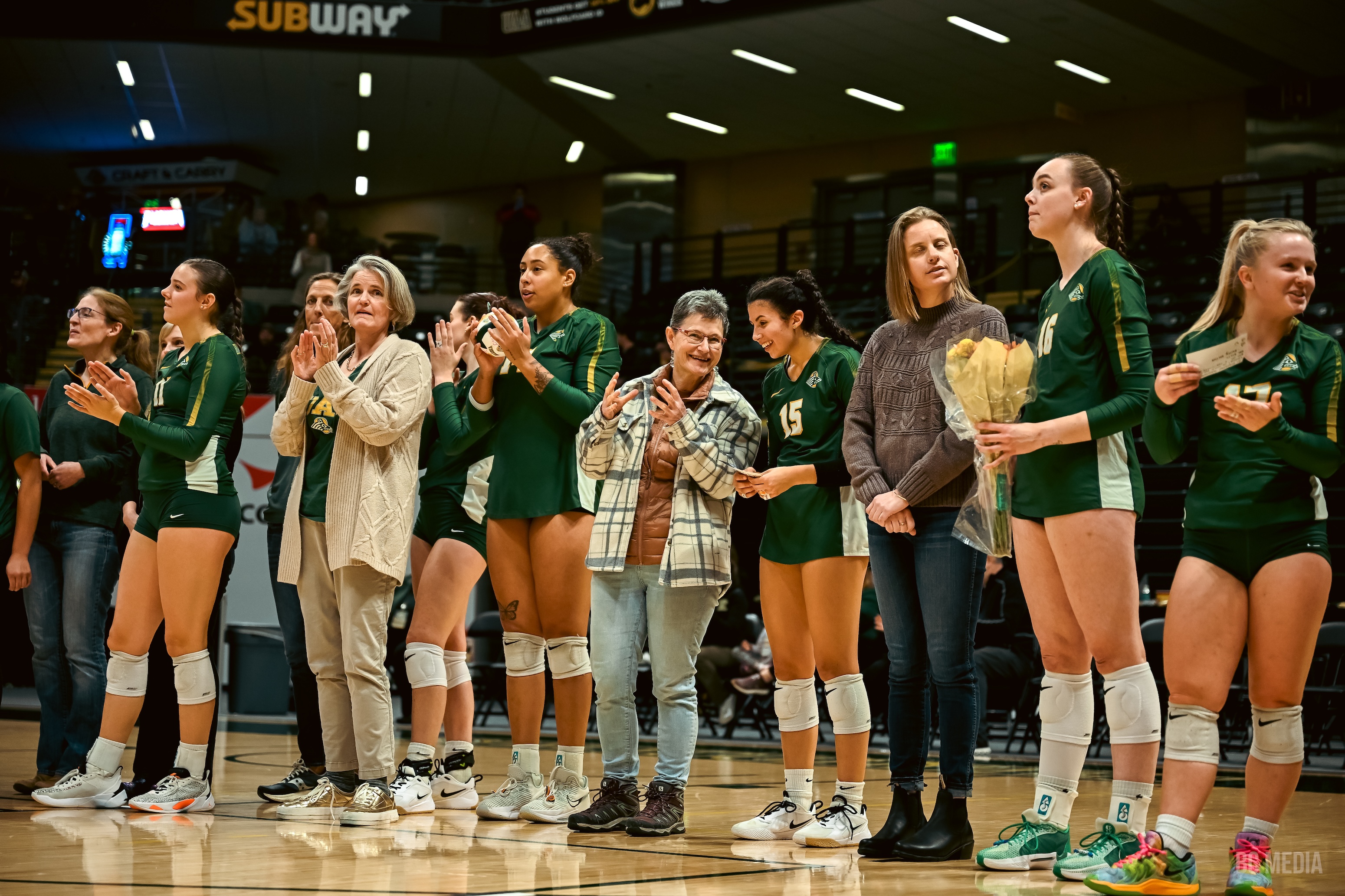 Volleyball athletes in green jerseys stand on the court next to faculty. 