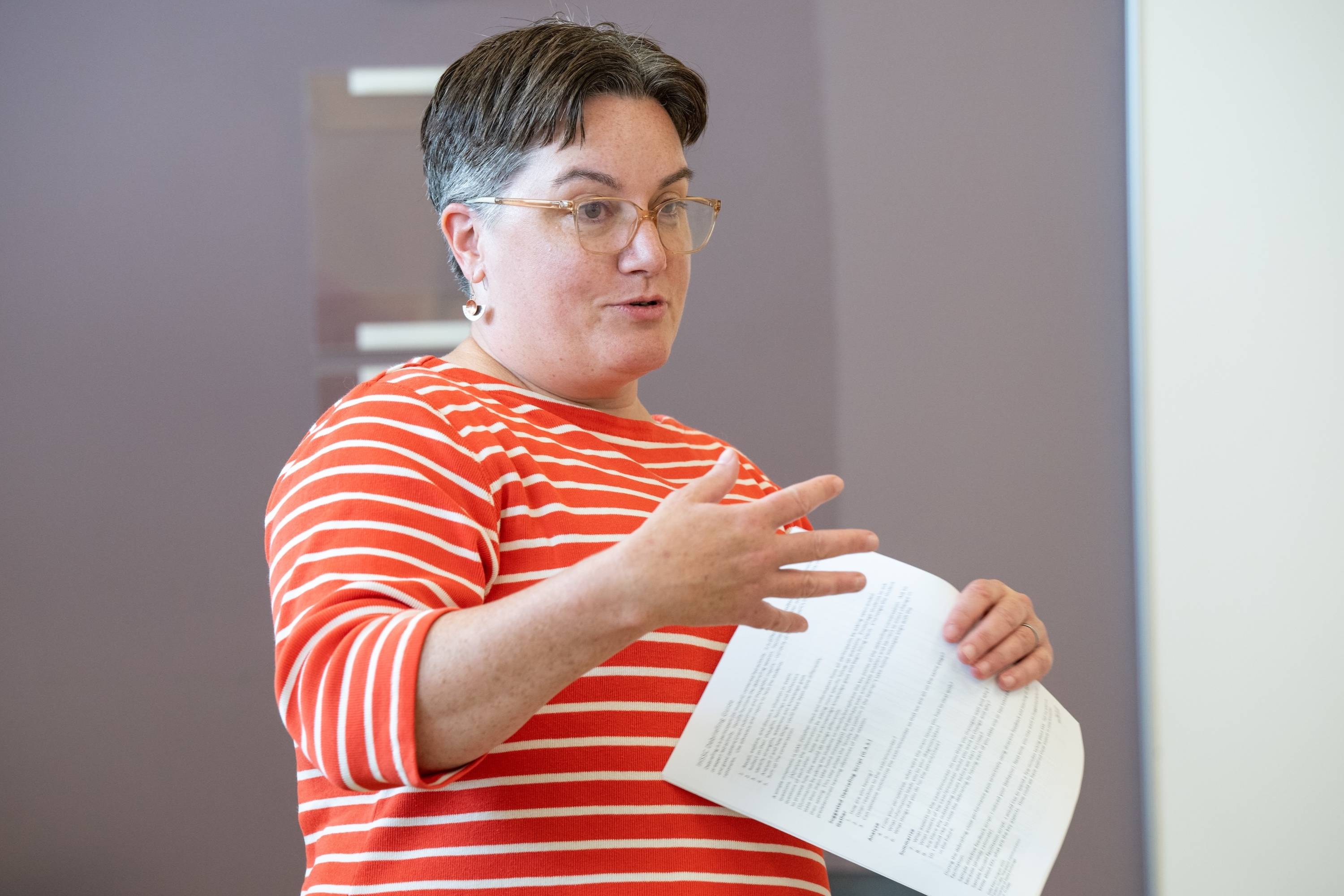 Sara Hannon, wearing a red and white horizontally striped shirt, stands indoors against a purple and white wall. She is holding a printed sheet of paper in one hand and gesturing with the other, as she presents or teaches. Sara's facial expression and body language indicate engagement, possibly in a classroom or meeting setting. 