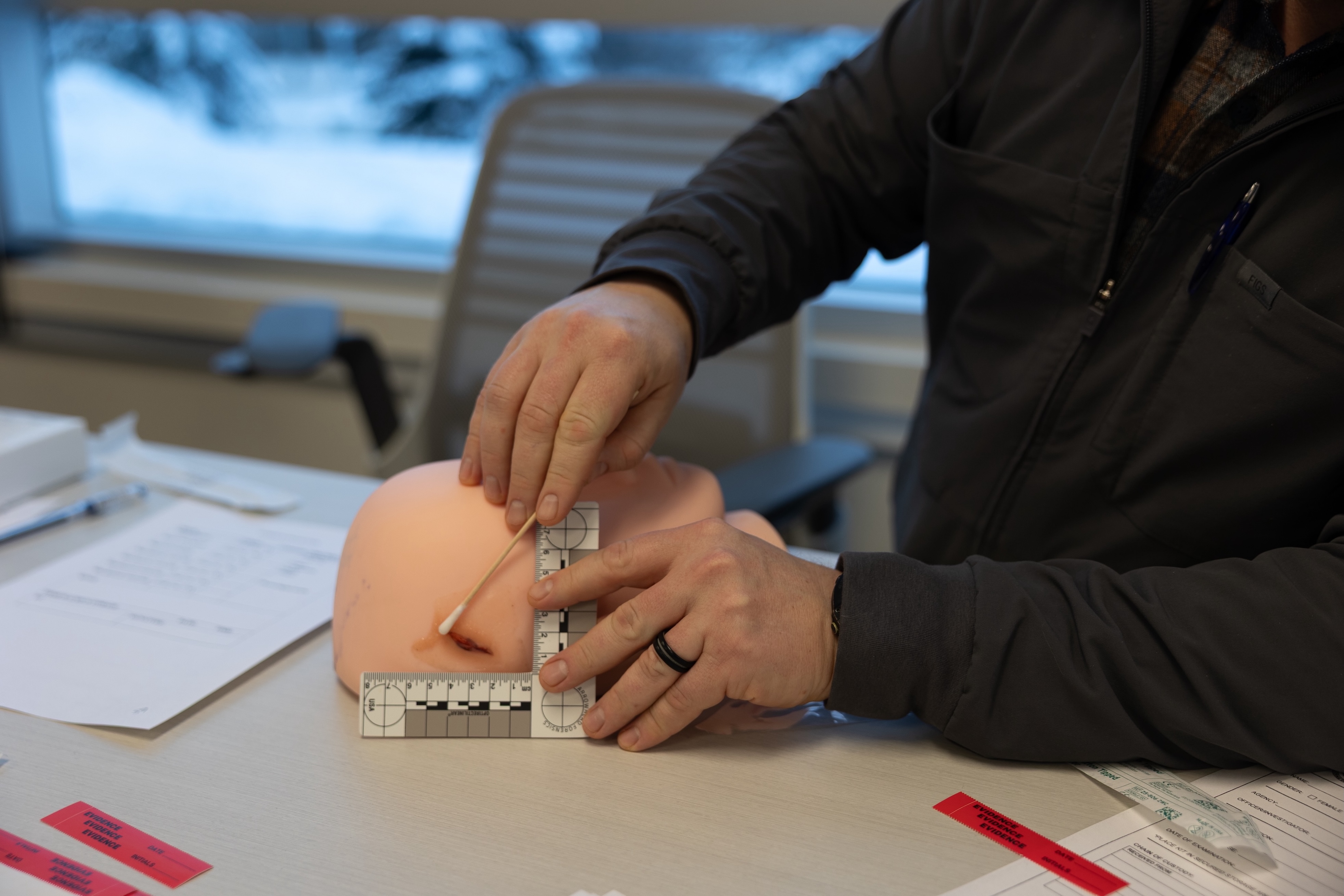 An ACFTA associate is picutured holding a mannequin head with a fake cut over the ear. He holds a square measuring tool next to the cut and pretends to swab it. 